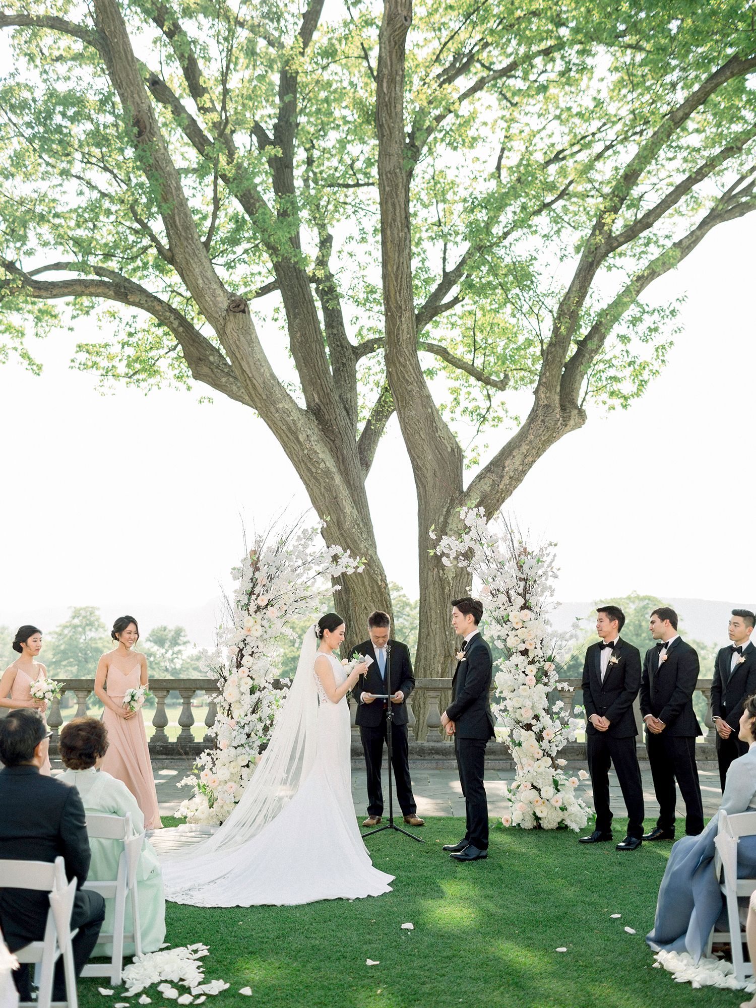 Outdoor wedding ceremony under large tree with white floral arrangements and seated guests.