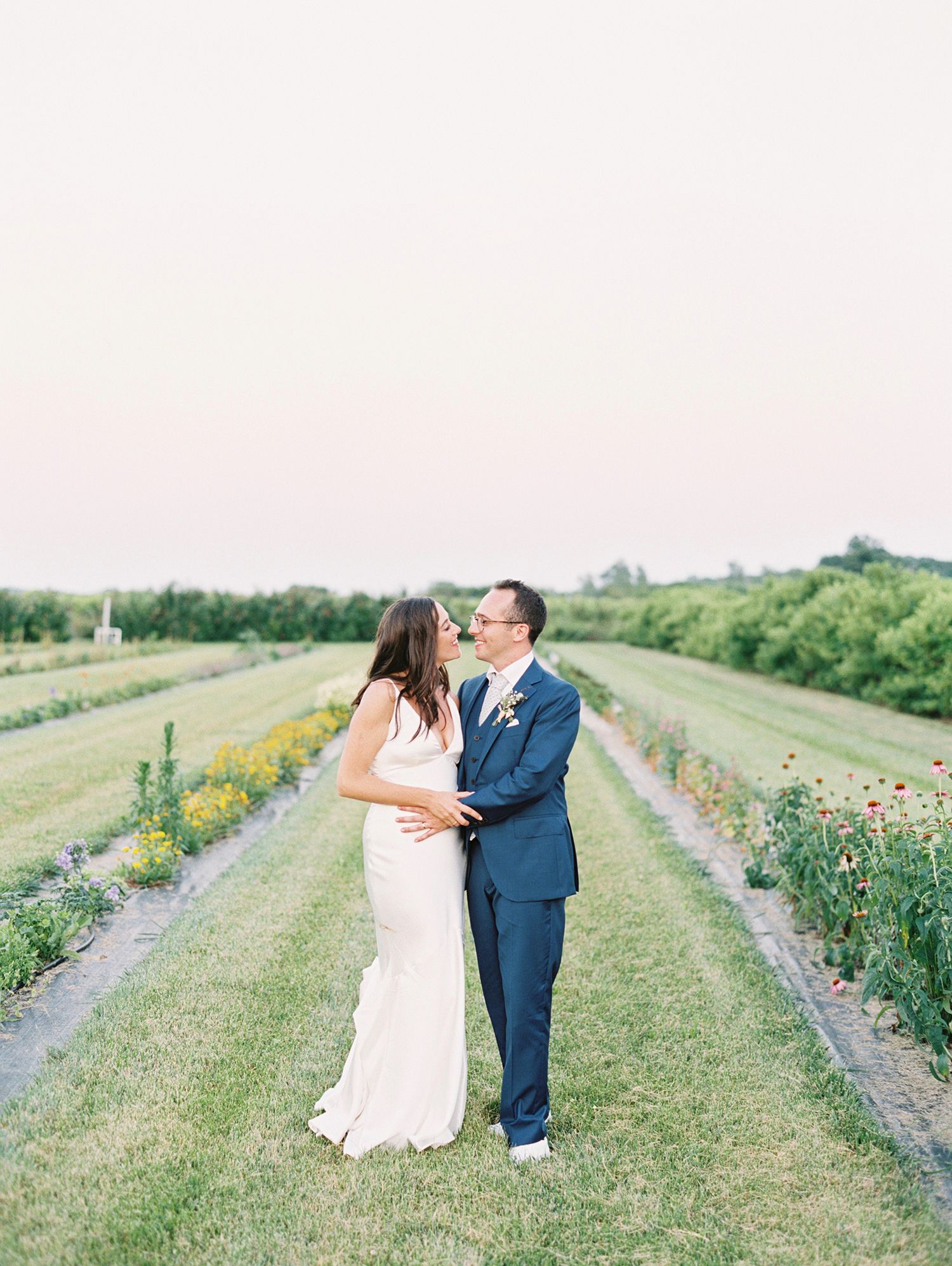 A romantic couple poses in a flower field wearing wedding attire against a bright summer landscape.