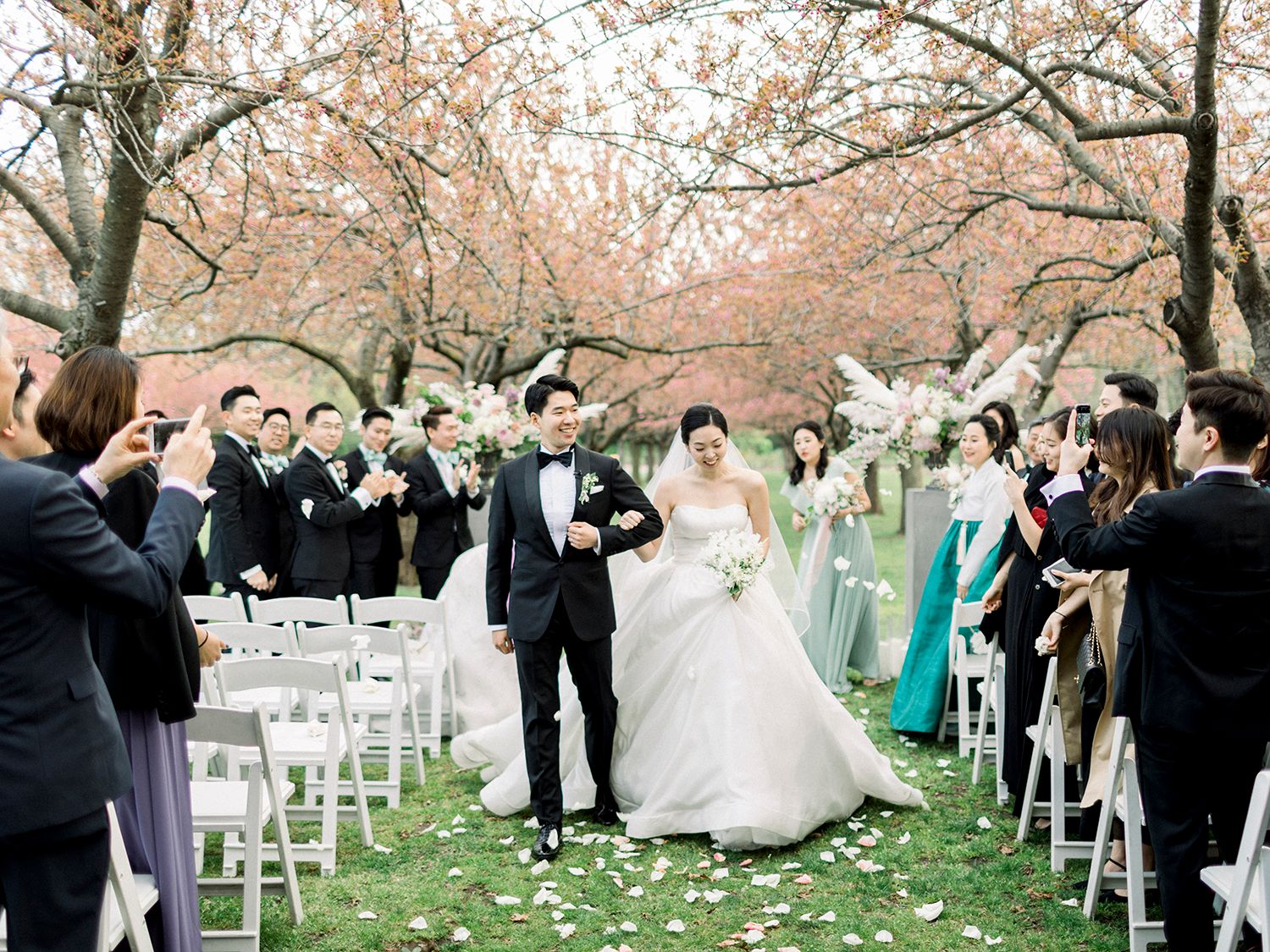 Wedding ceremony in progress under flowering cherry trees with guests watching.