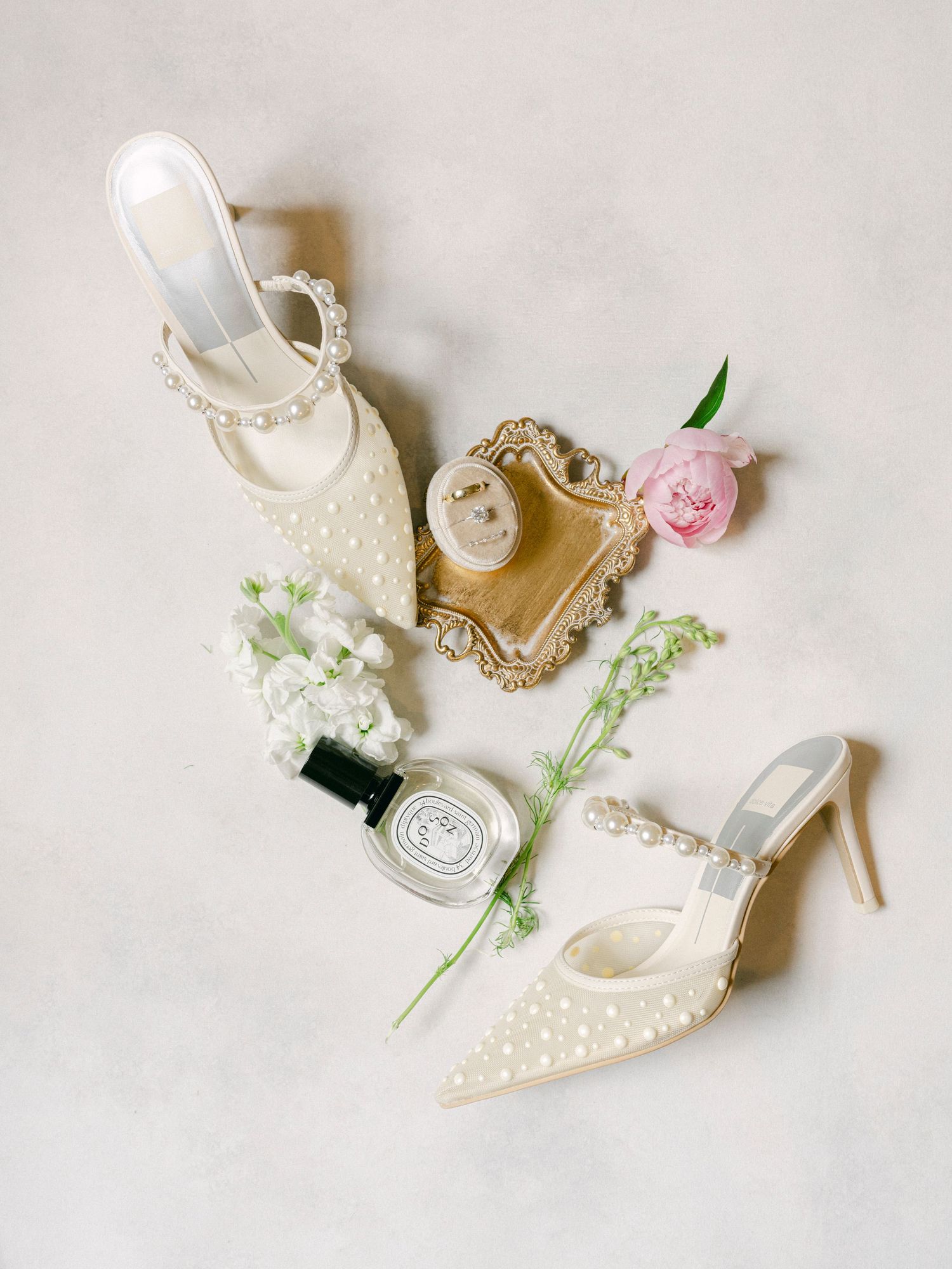 Delicate white bridal shoes with pearl details arranged with small pink flowers on light background.