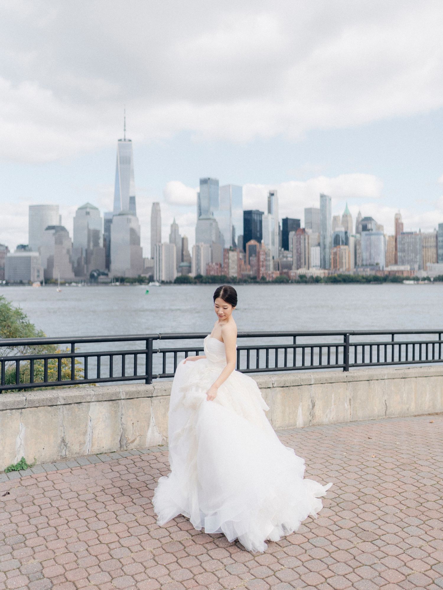 A bride in a flowing white dress poses against the New York City skyline backdrop along the waterfront.