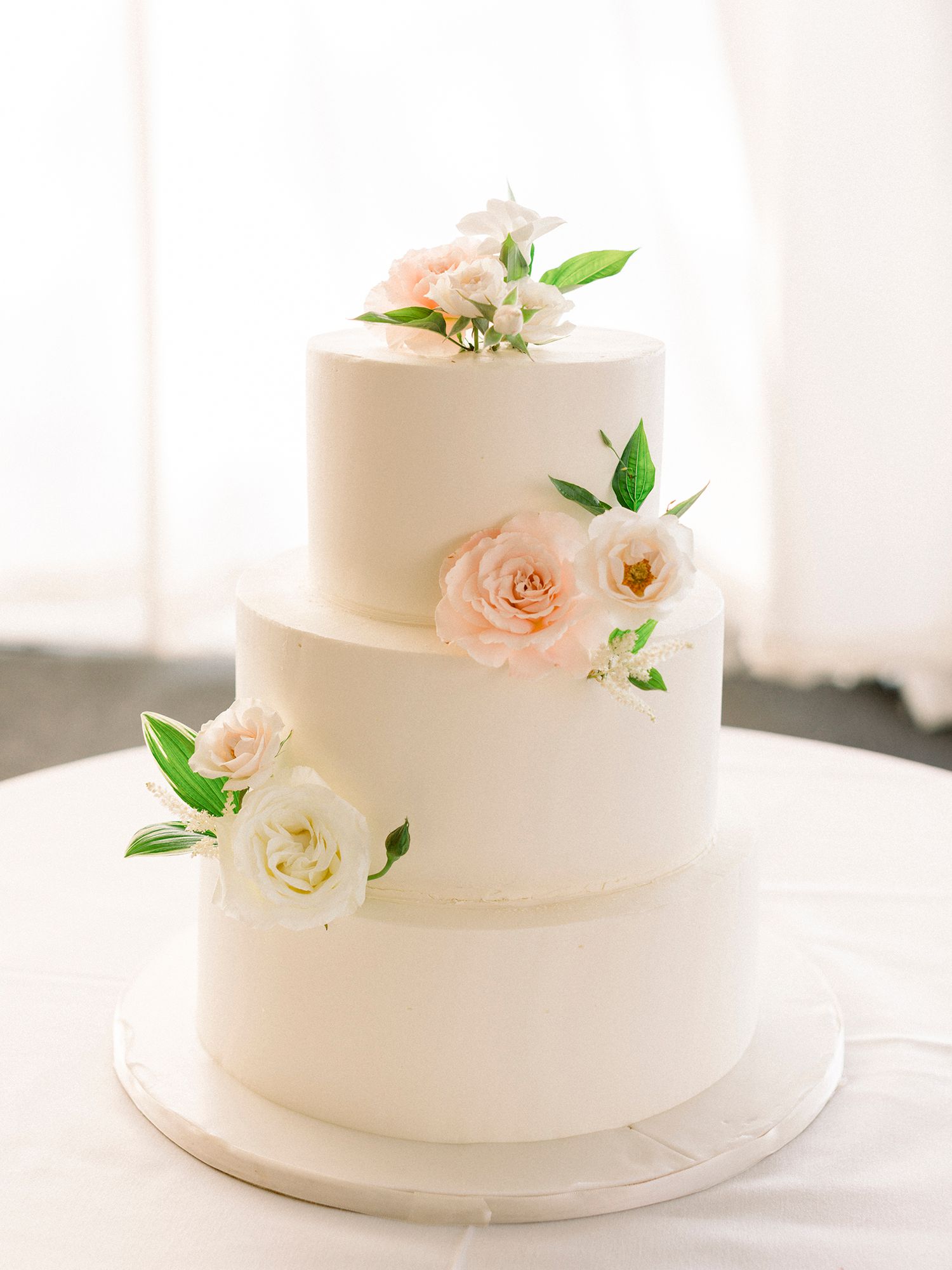 Minimalist white wedding cake decorated with soft pink roses and green foliage.