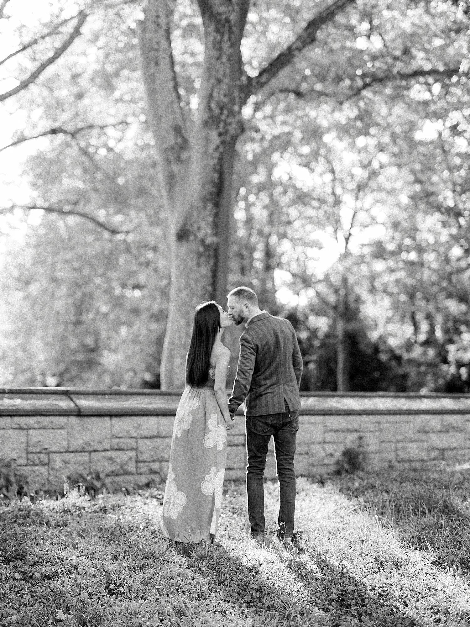 A couple walks together on a grassy path surrounded by trees in black and white.