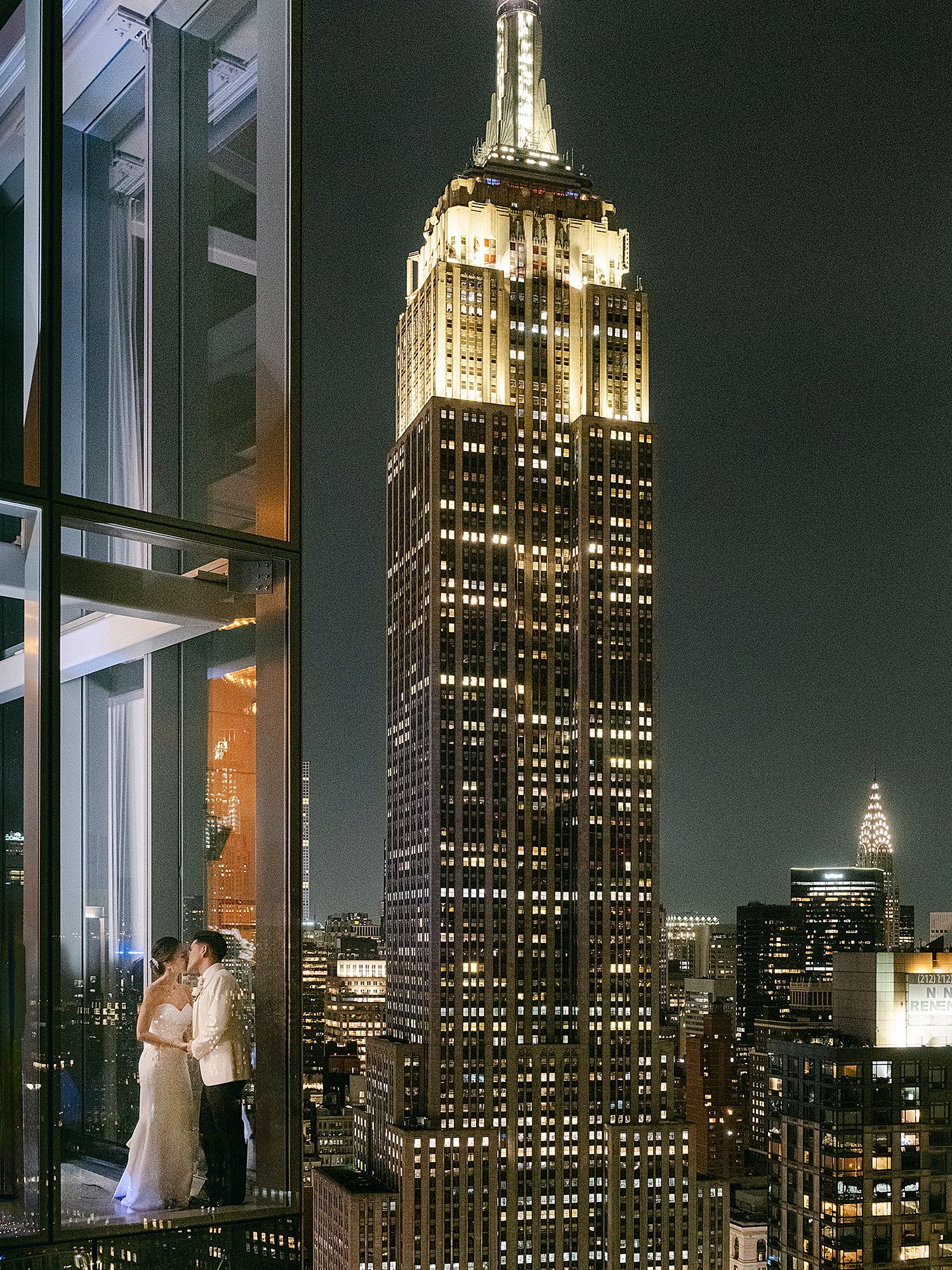 Wedding dress silhouetted against illuminated Empire State Building at night through large windows. NYC Wedding Photographer