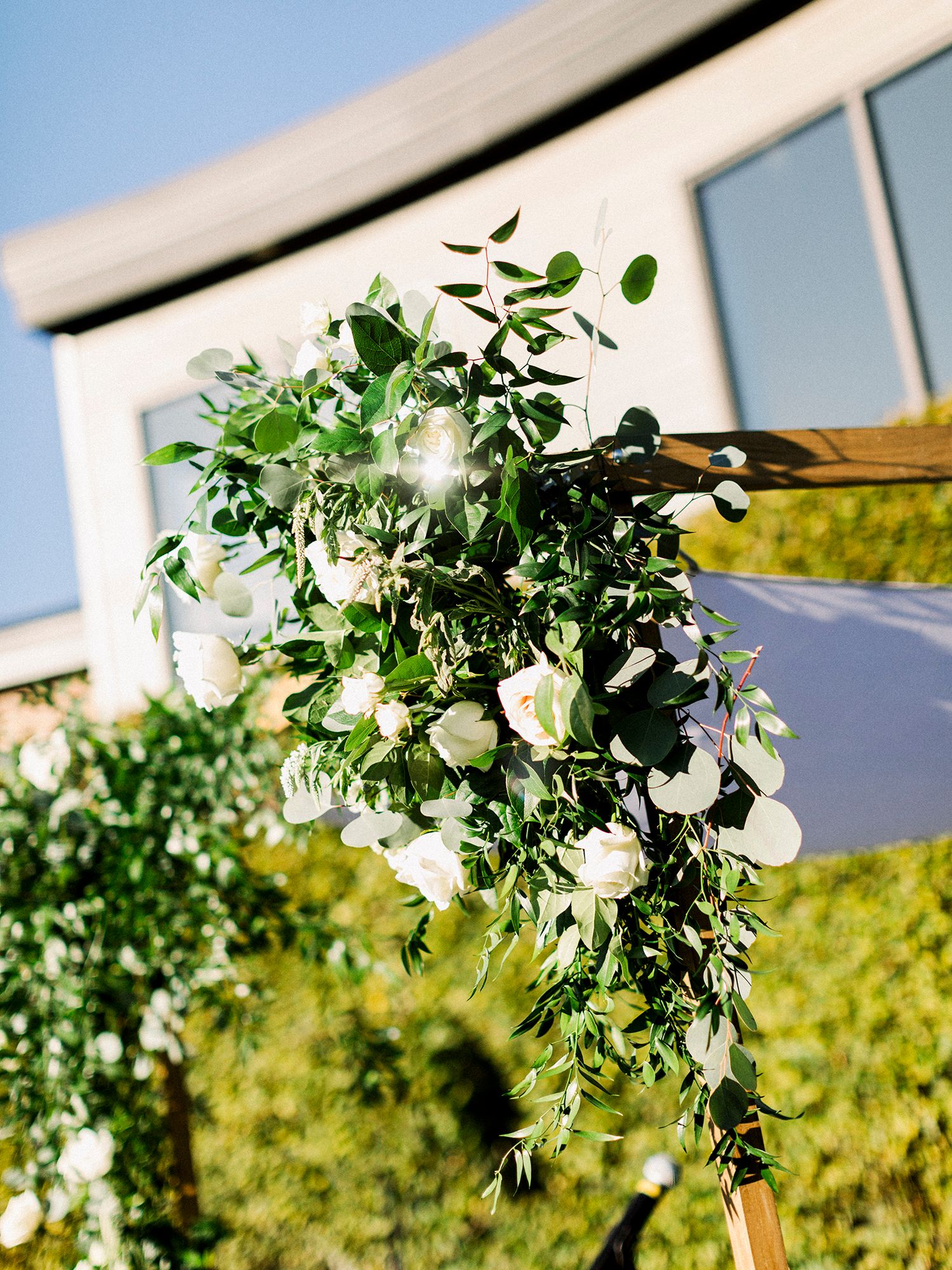 Floral arch with white roses and greenery against blue sky at outdoor wedding venue.