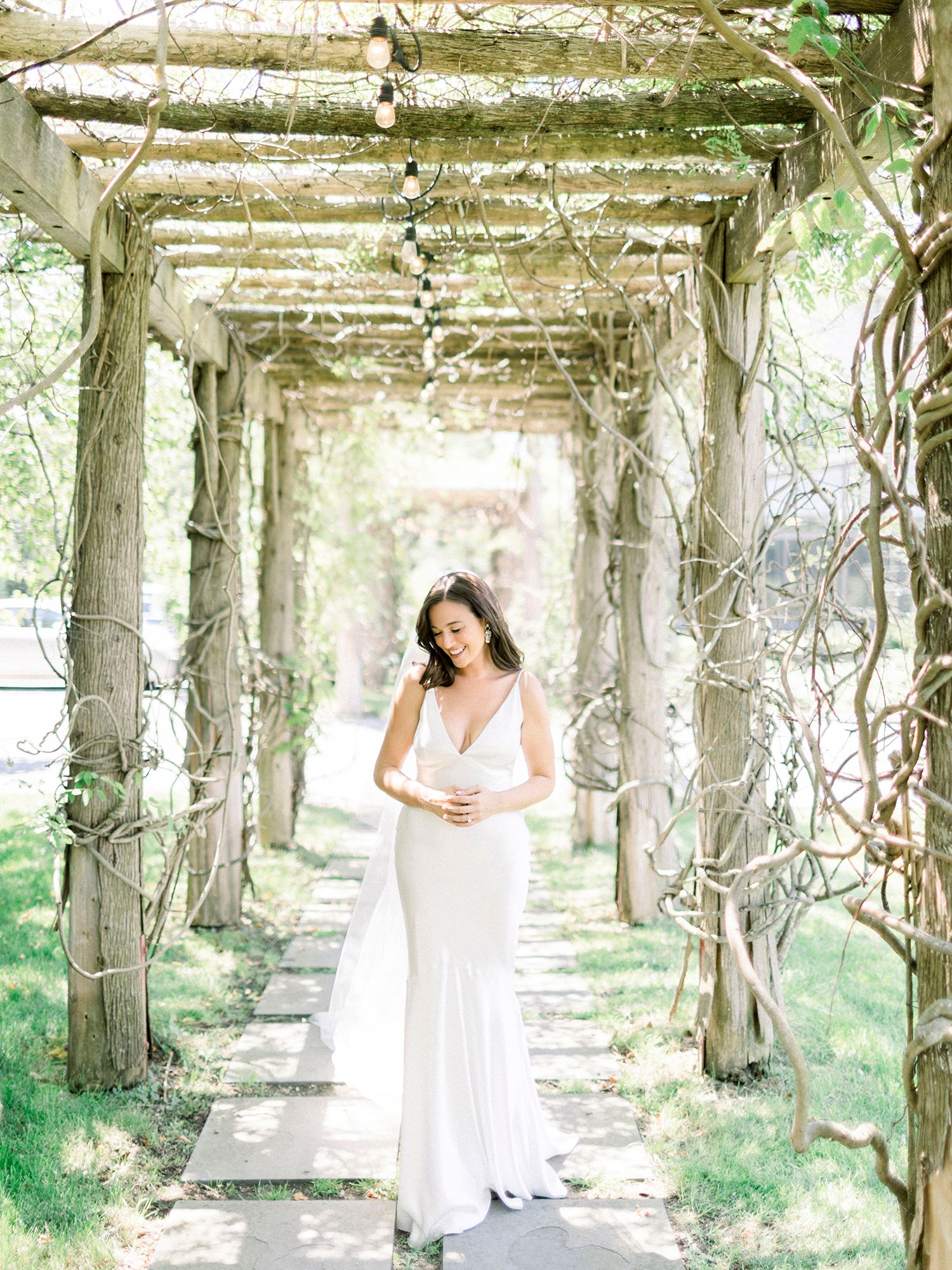 Bride in white dress posing under a rustic wooden pergola covered in vines.