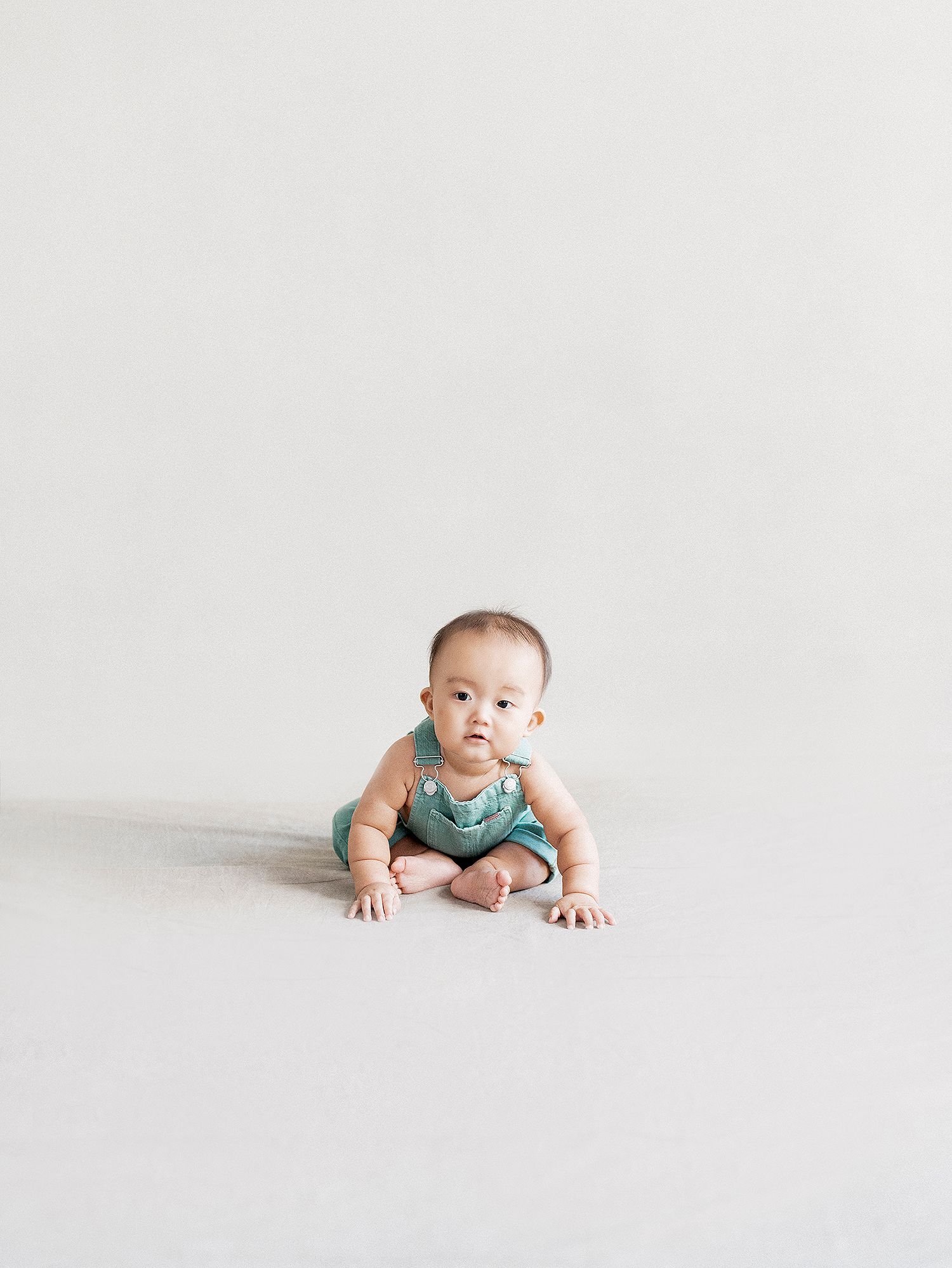 Baby in teal onesie crawls on white studio floor while looking at camera with curious expression.