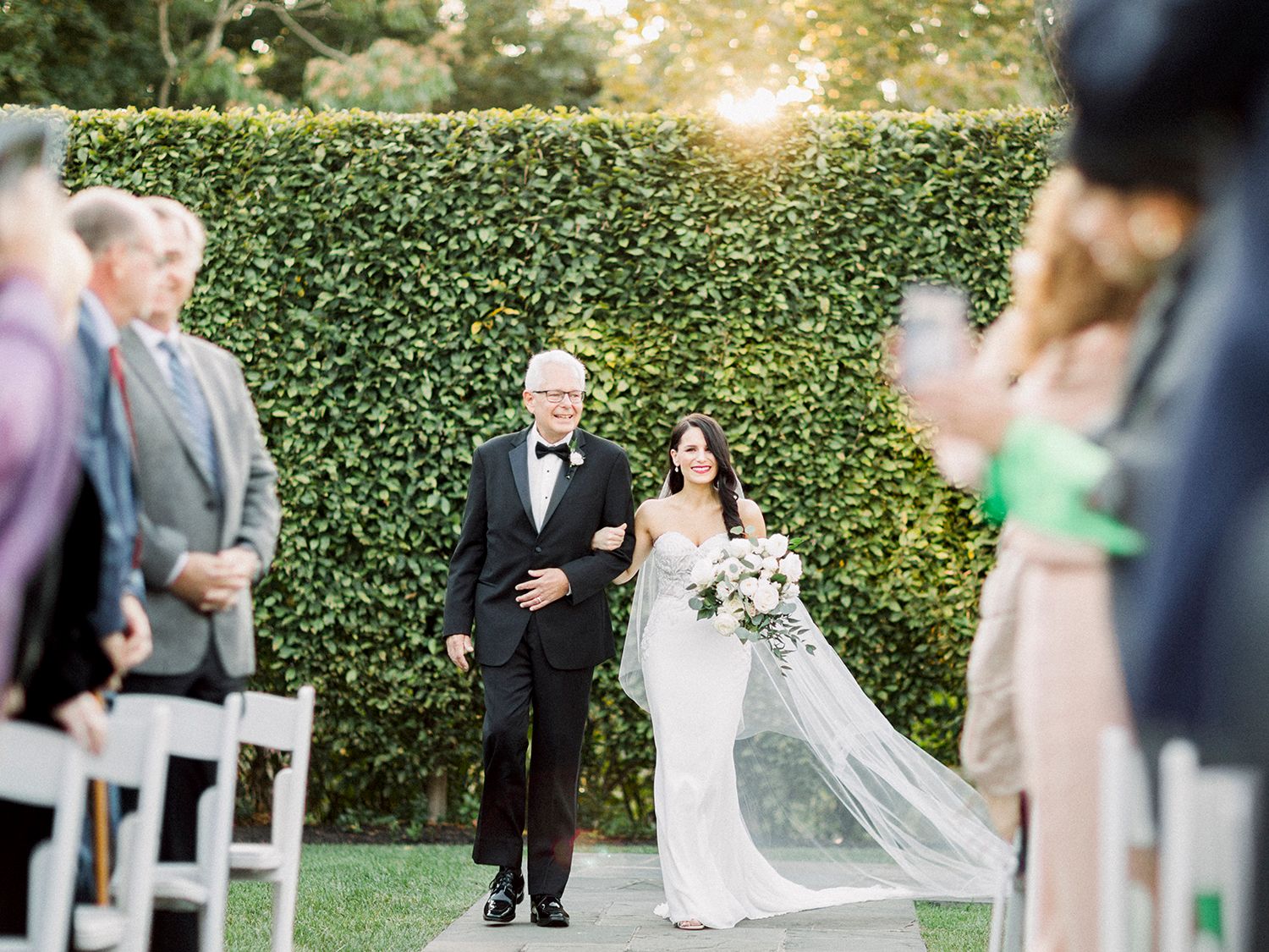 Elegant outdoor wedding ceremony with guests seated in white chairs surrounded by lush greenery.