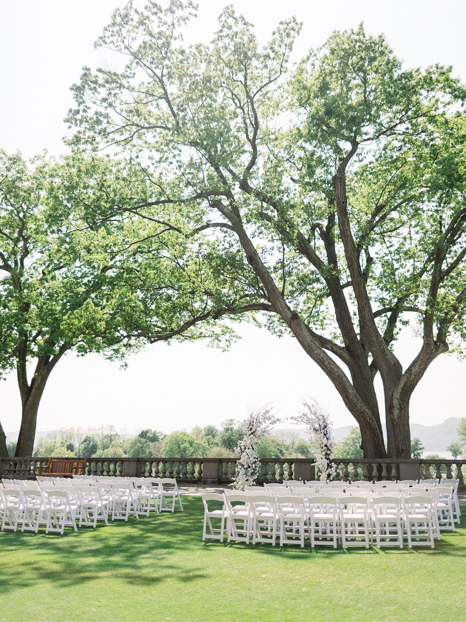 Outdoor wedding ceremony setup with white chairs arranged on a lush green lawn under mature trees.