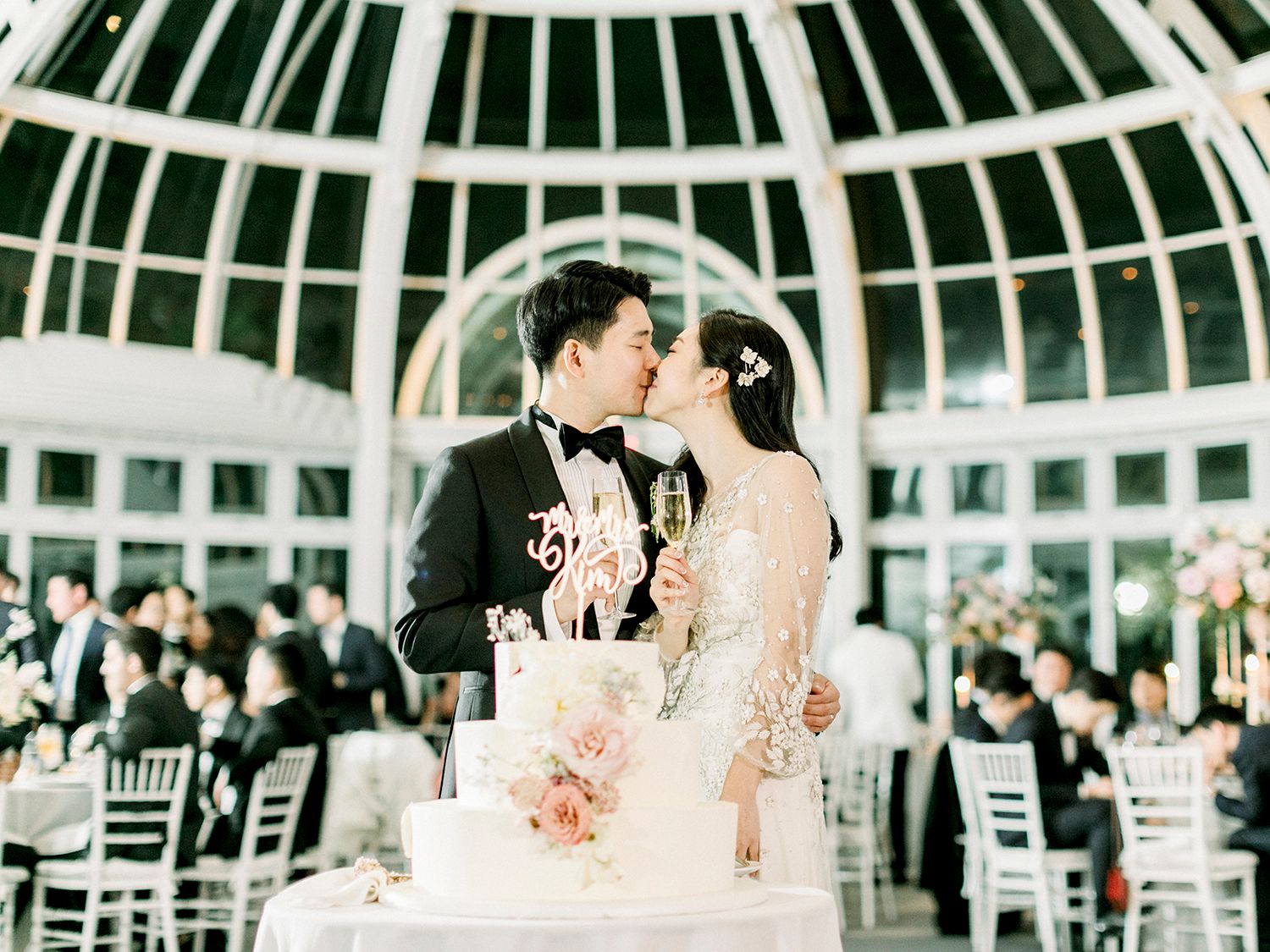 Elegant wedding reception setup under a grand glass dome conservatory with crystal chandeliers and white decor.