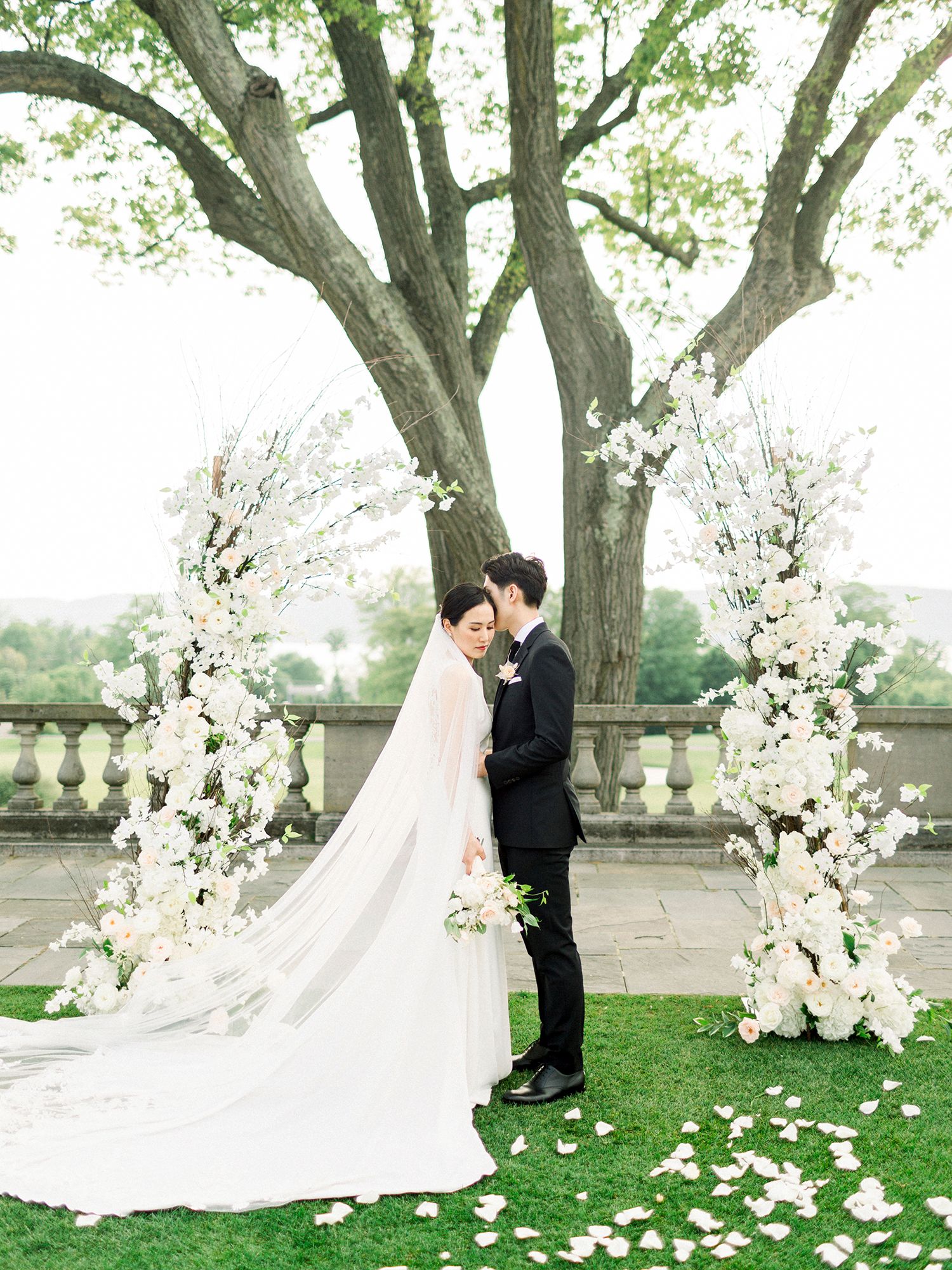 Elegant wedding ceremony setup outdoors with white floral arrangements and trees creating a romantic backdrop. NYC wedding photographer.