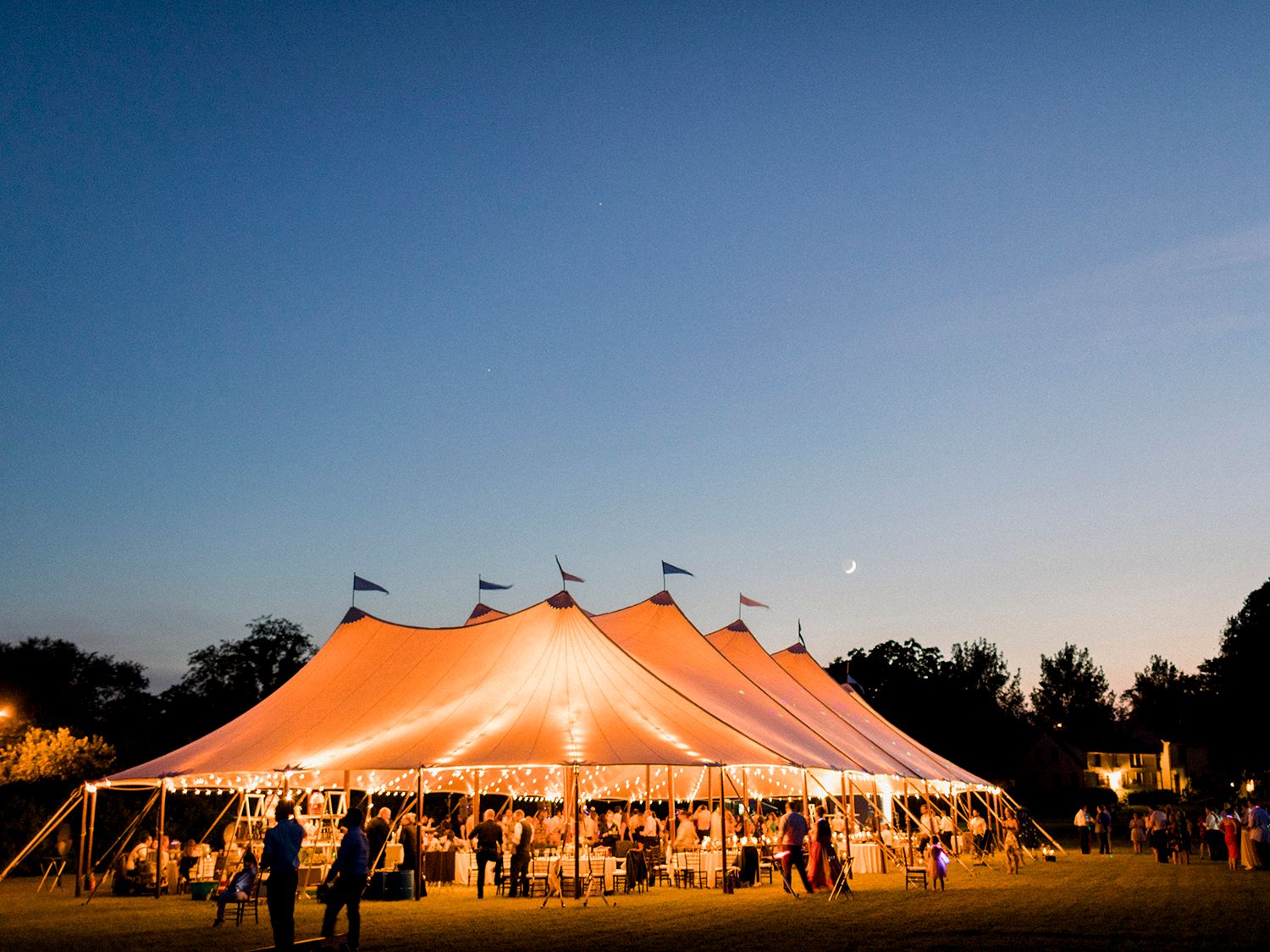 Large orange wedding tent illuminated at dusk with guests gathering outside.