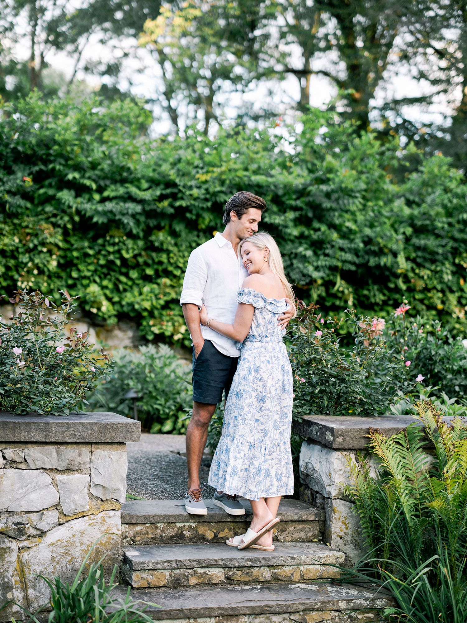 A romantic couple sits together on stone garden steps surrounded by lush greenery and flowers.