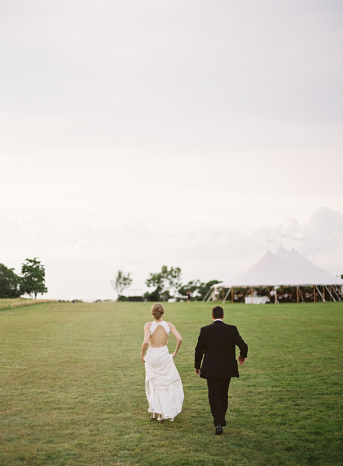 Bride and groom walk across an open green field at sunset.