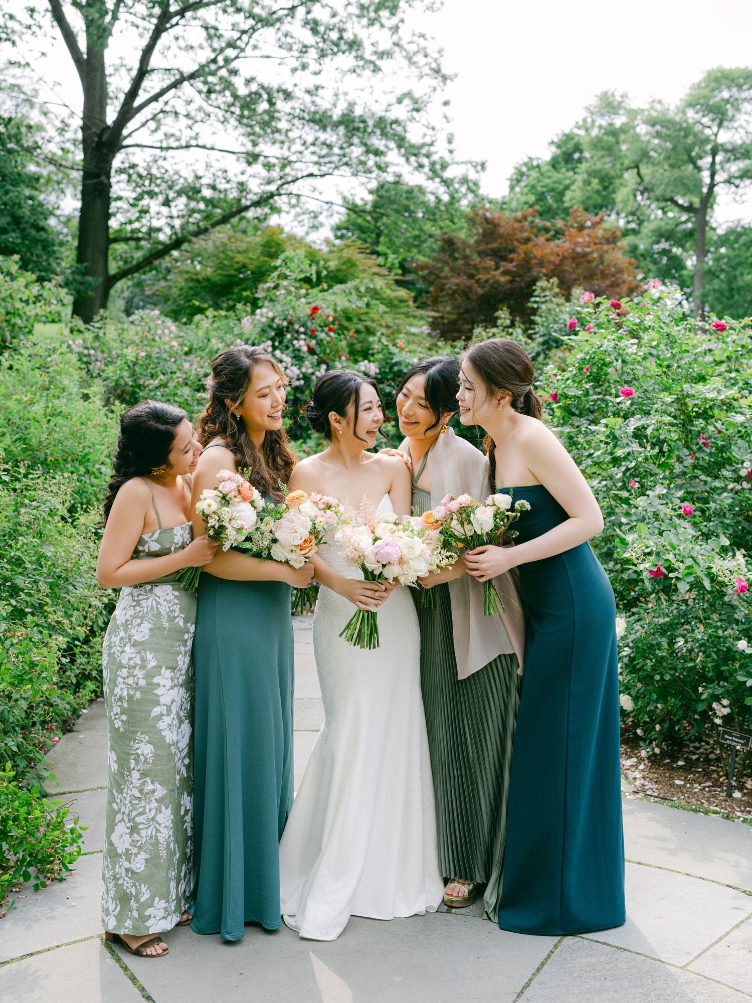 A group in elegant dresses gather in a garden holding floral bouquets.