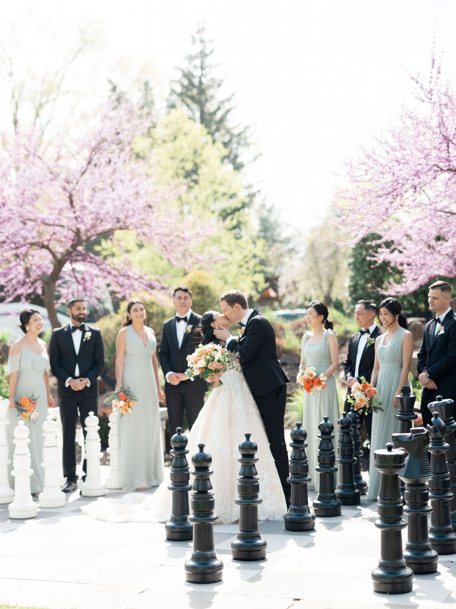 Wedding party lined up between large decorative chess pieces outdoors under cherry blossoms.