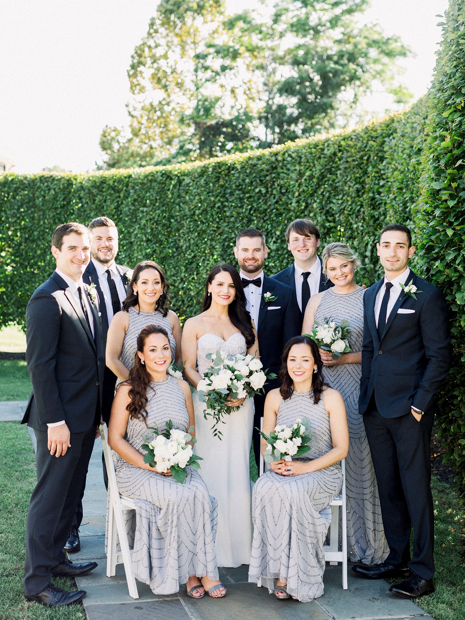 Wedding party poses together in silver dresses and black tuxedos against garden hedge.
