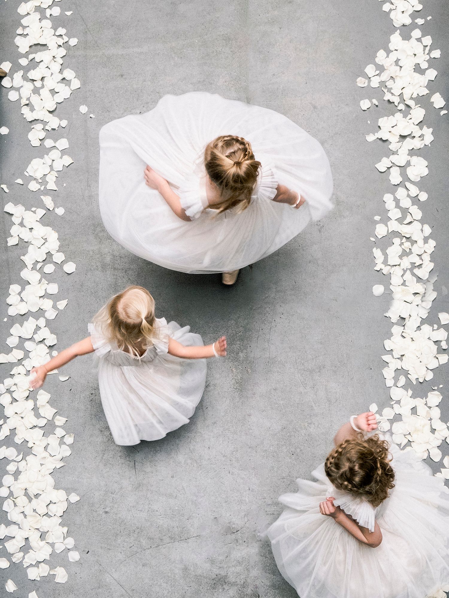 Overhead view of flower girls in white dresses twirling on a grey floor with floral decorations. NYC wedding photographer.