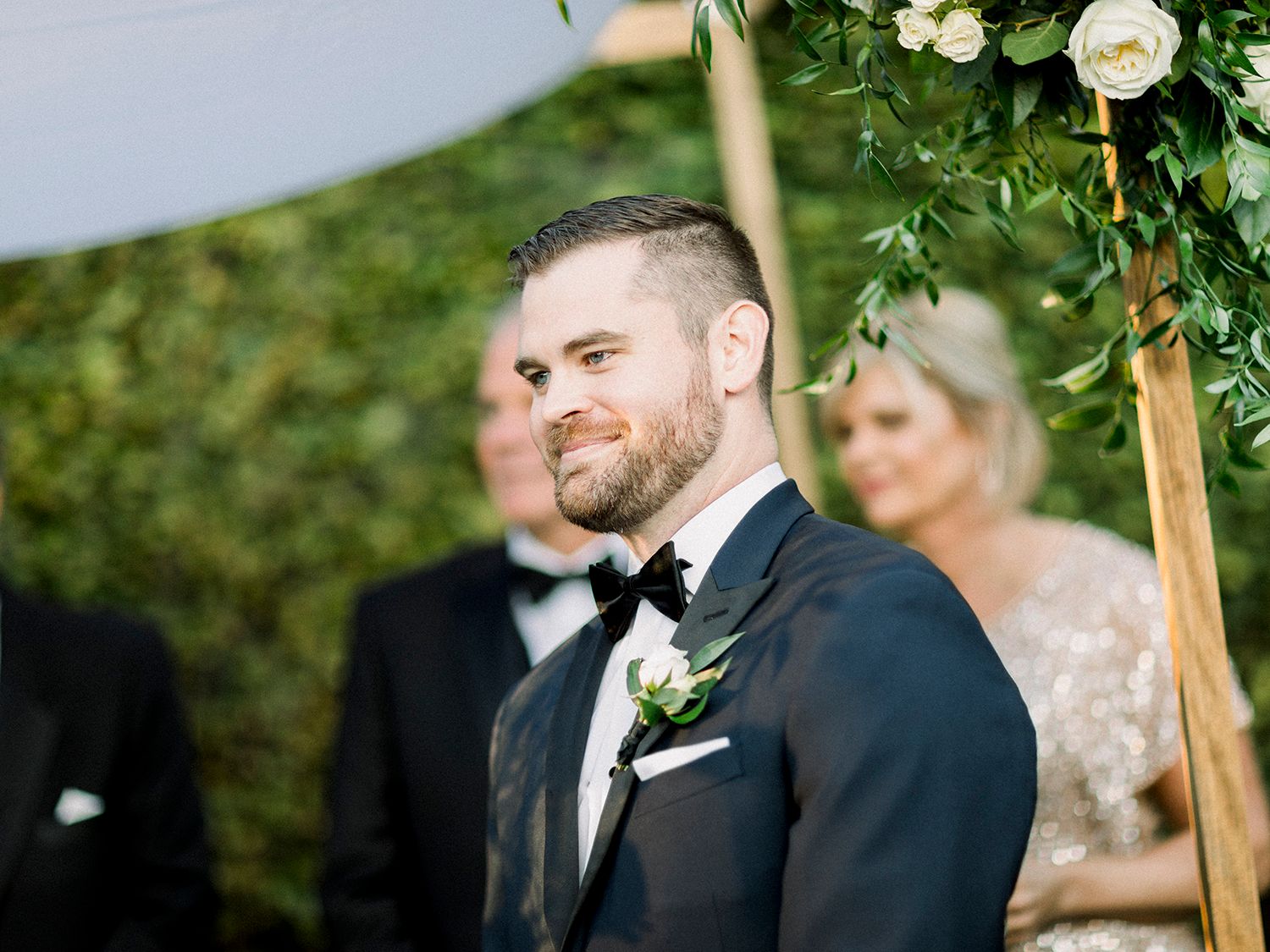 Wedding ceremony participants stand beneath floral arch during outdoor celebration.