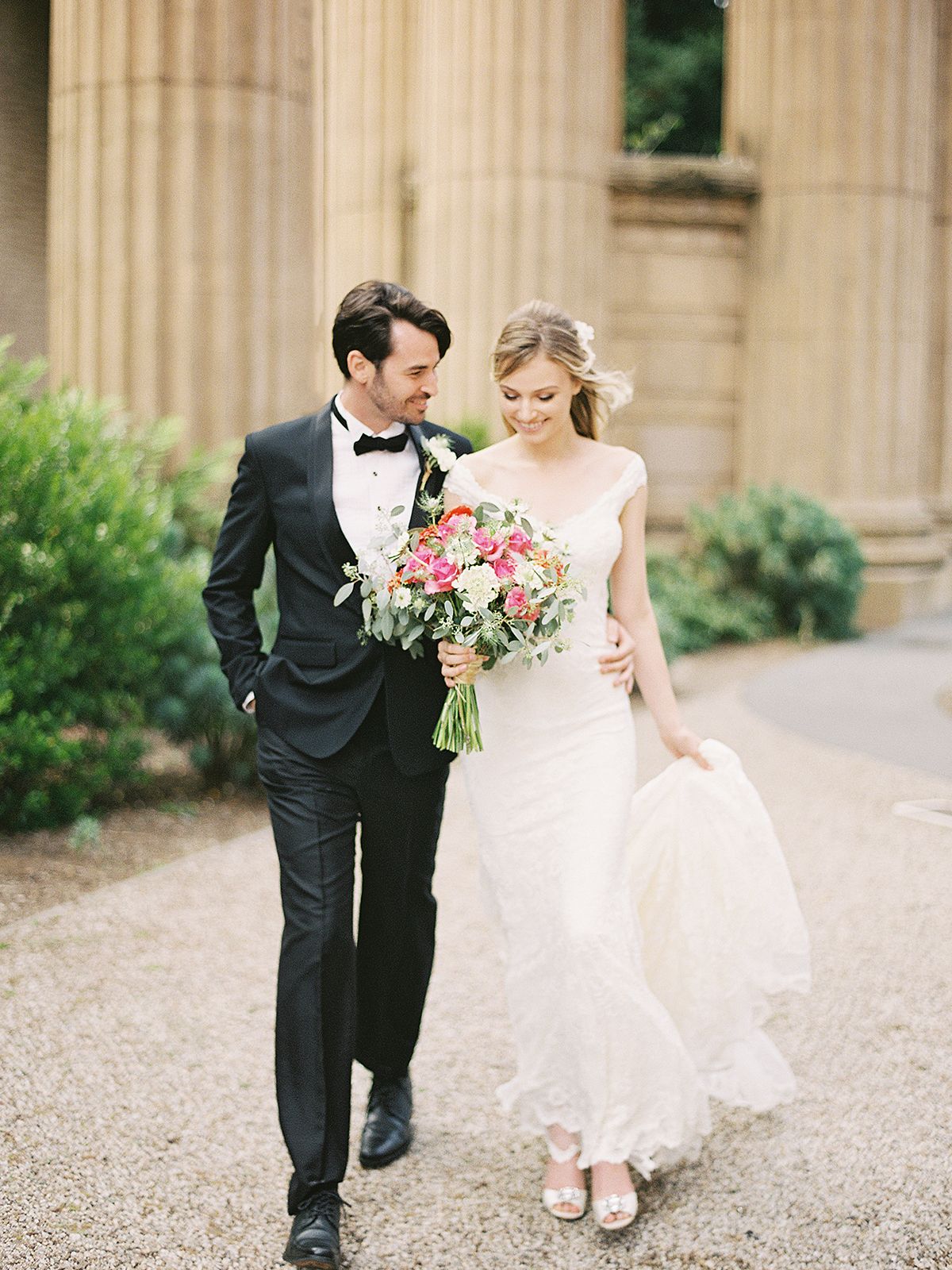 Couple walks among classical stone columns in formal wedding attire.