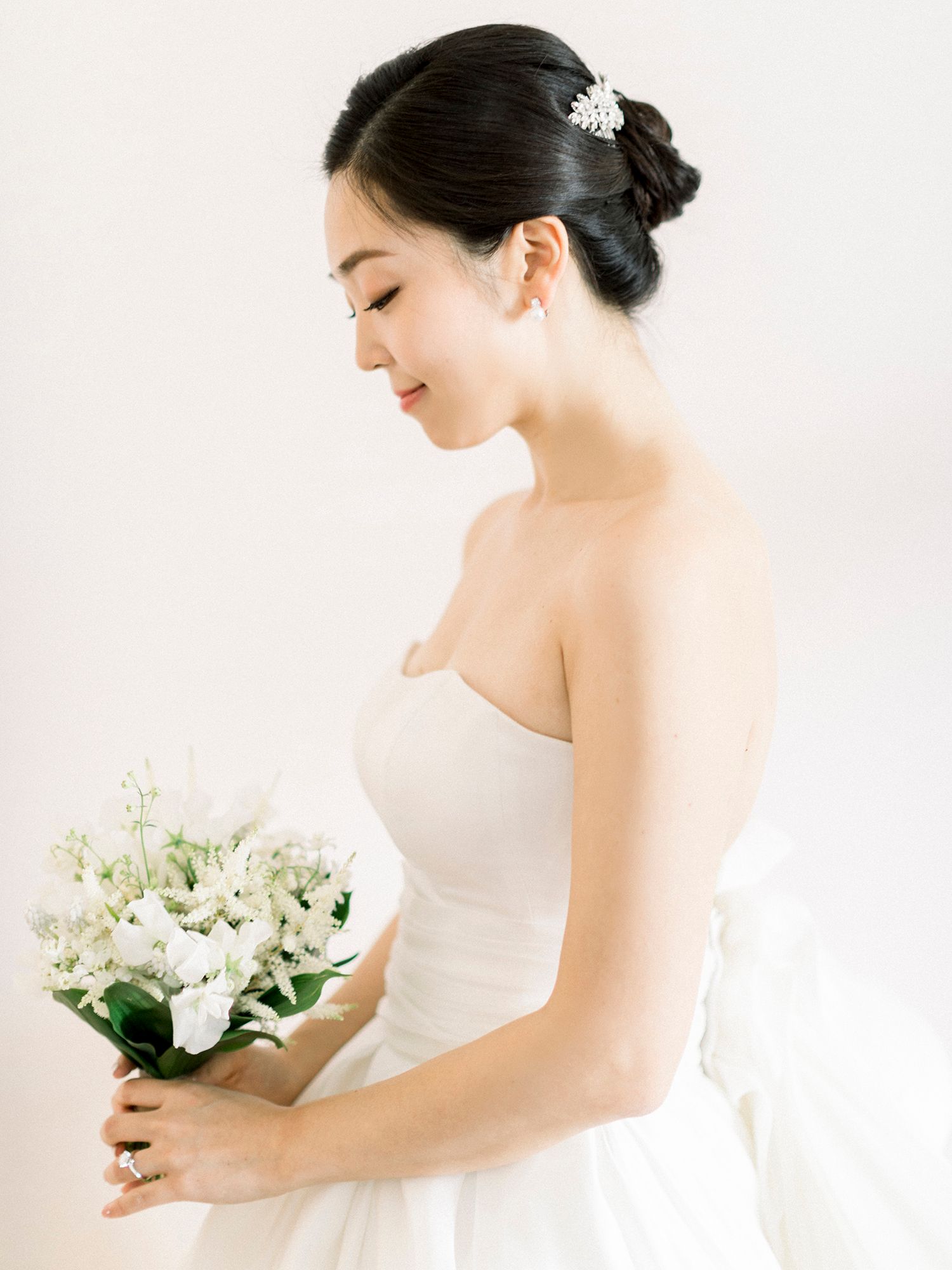 Elegant bride in white dress holds a delicate bouquet of white flowers against a bright background.