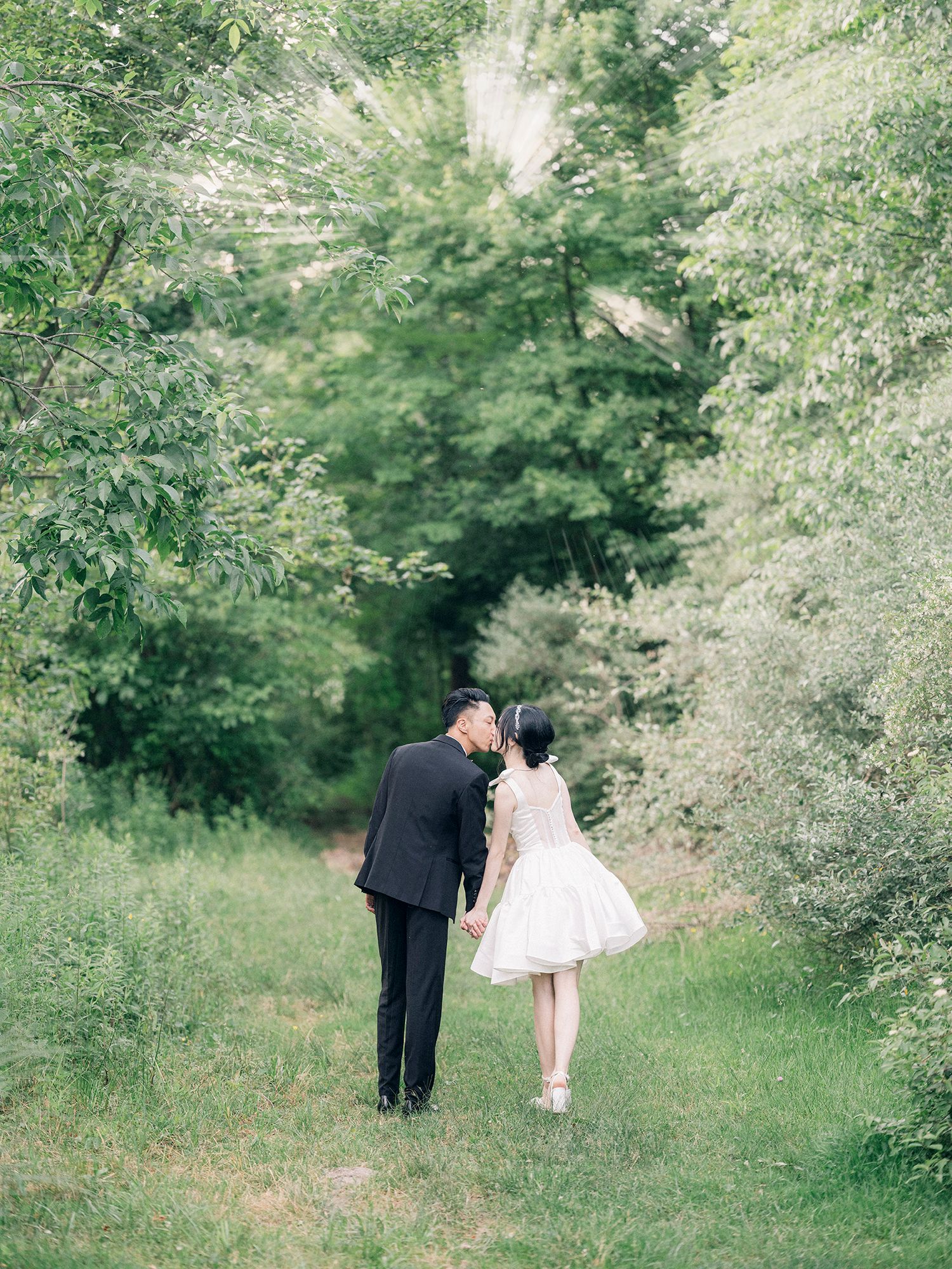 A couple walks along a lush green garden path surrounded by blooming trees and foliage. NYC wedding photographer.