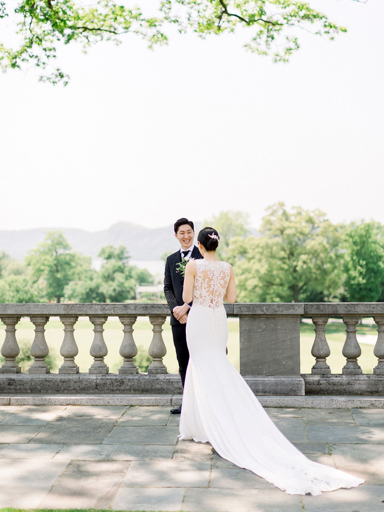 Elegant wedding couple poses on a stone balcony with mountain views and spring foliage overhead.