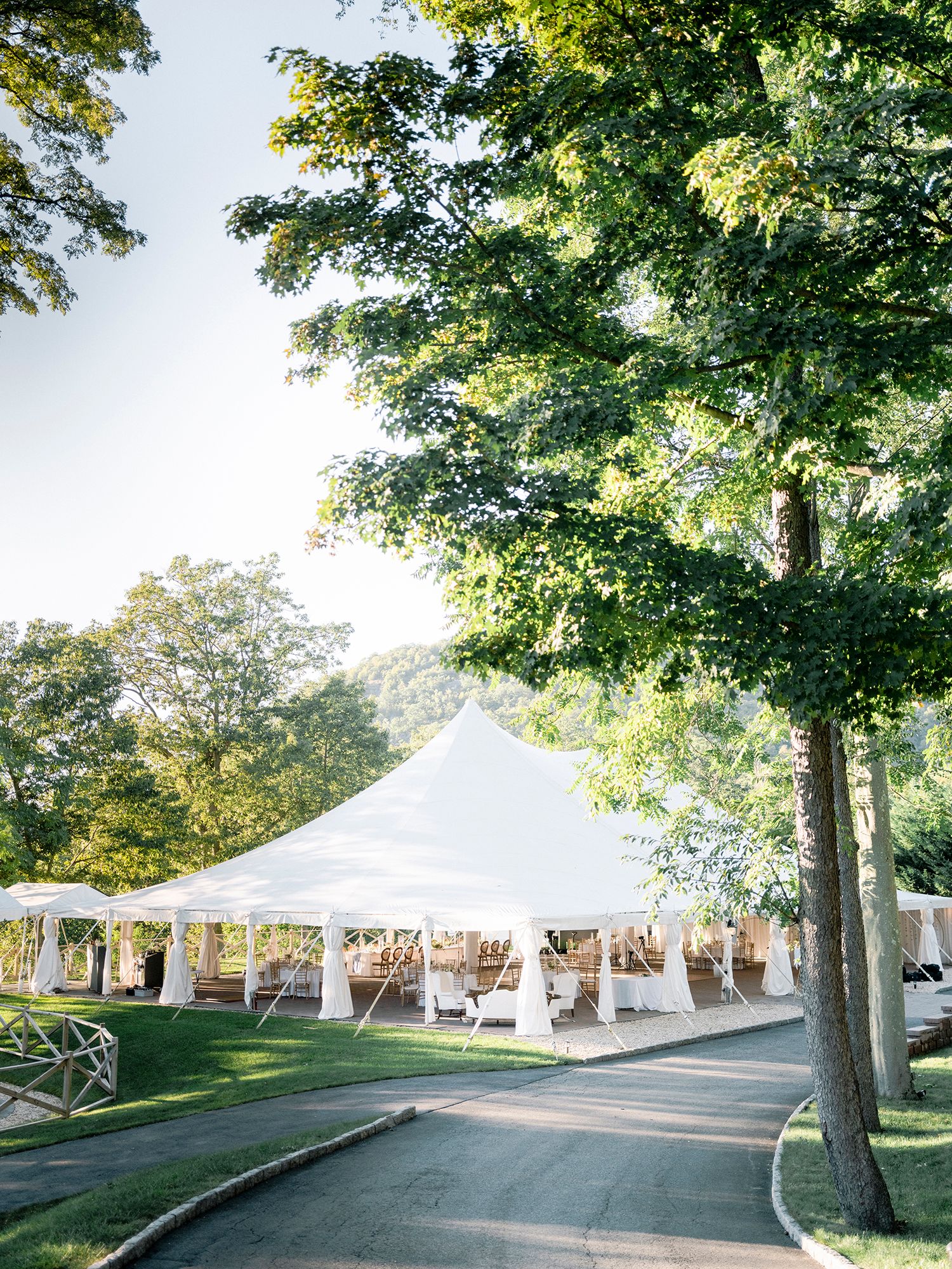White wedding tents line a winding paved path under green trees in an elegant outdoor venue setting.