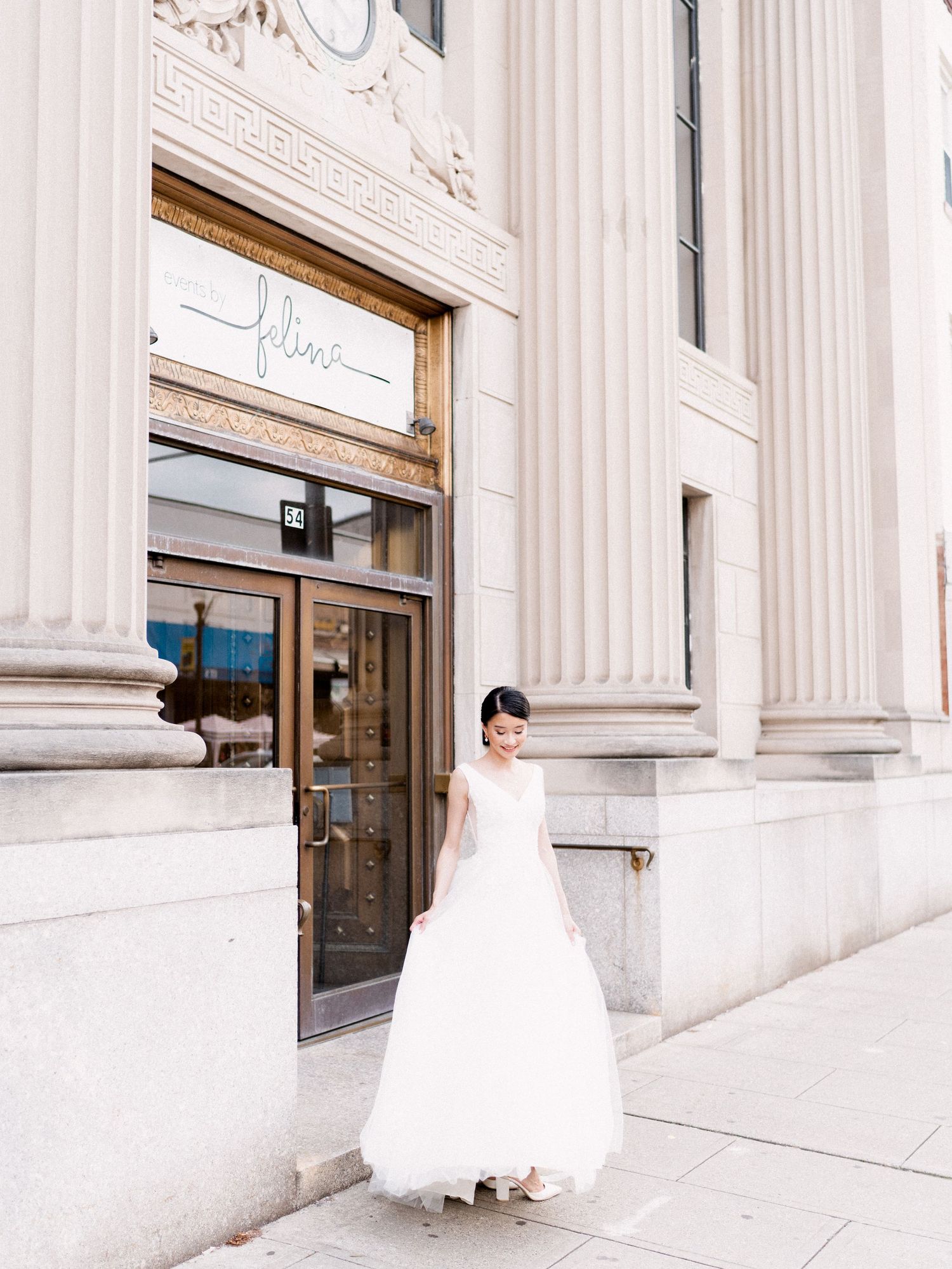 A bride in a white gown poses elegantly between classical columns of a grand building.