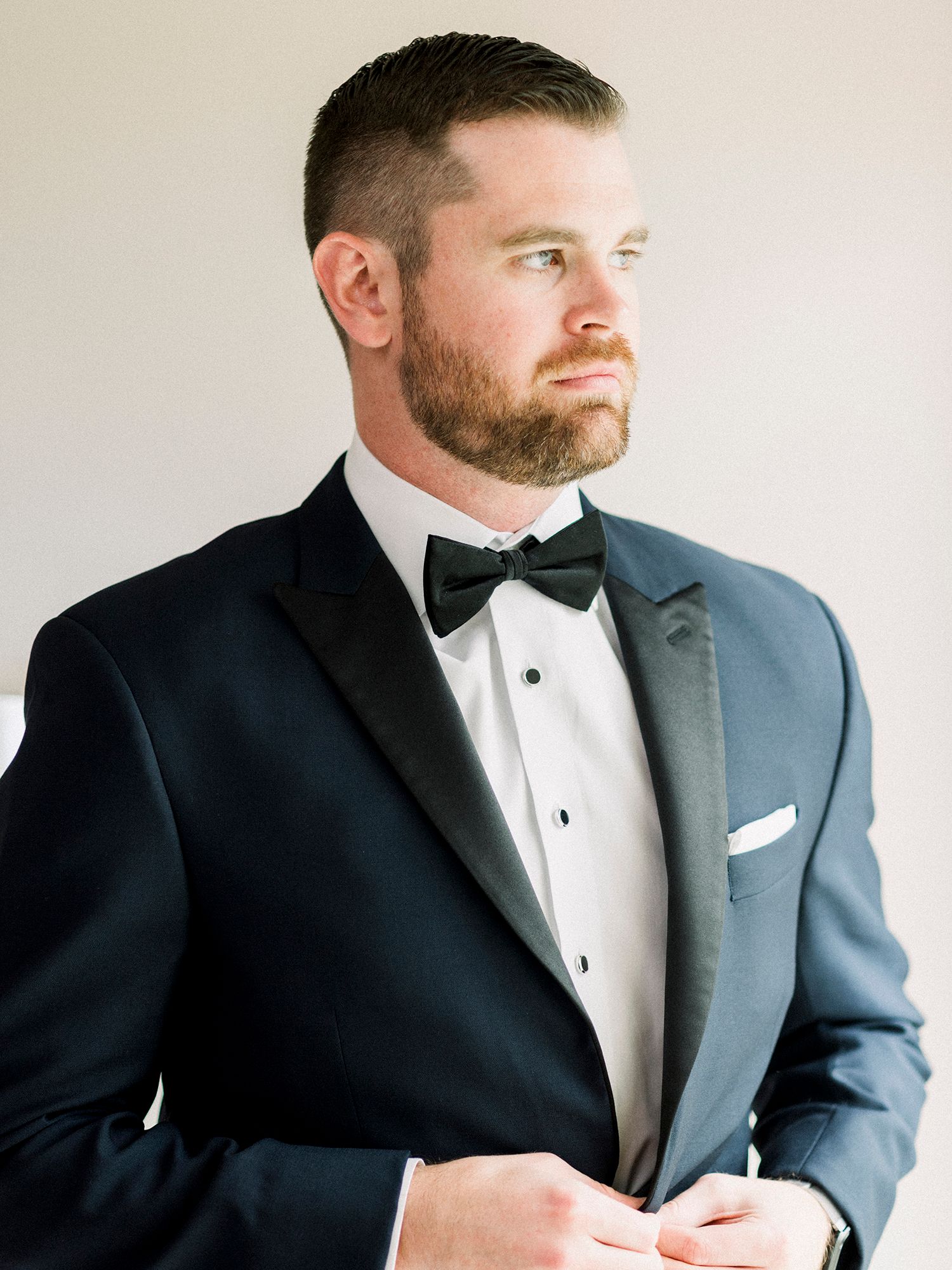 Man in formal tuxedo and bowtie poses against light background.