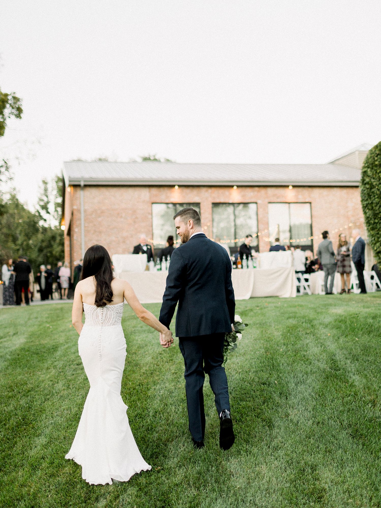 Wedding couple walking across a green lawn with brick building in the background.