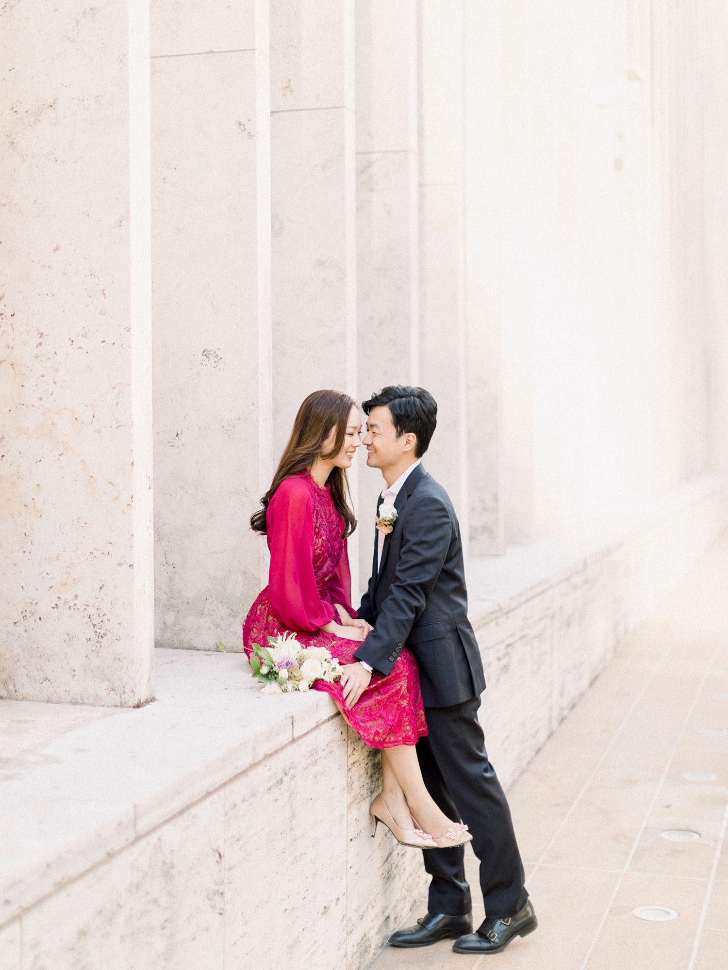 Elegant engagement portrait against white stone columns with red dress detail.