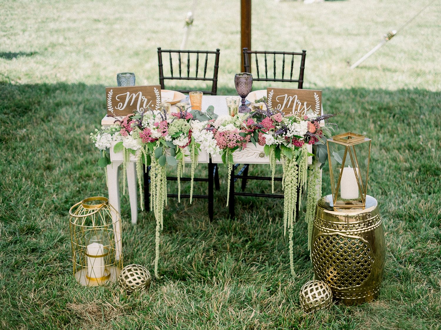 Elegant outdoor wedding reception setup with gold accents and chiavari chairs on green lawn.