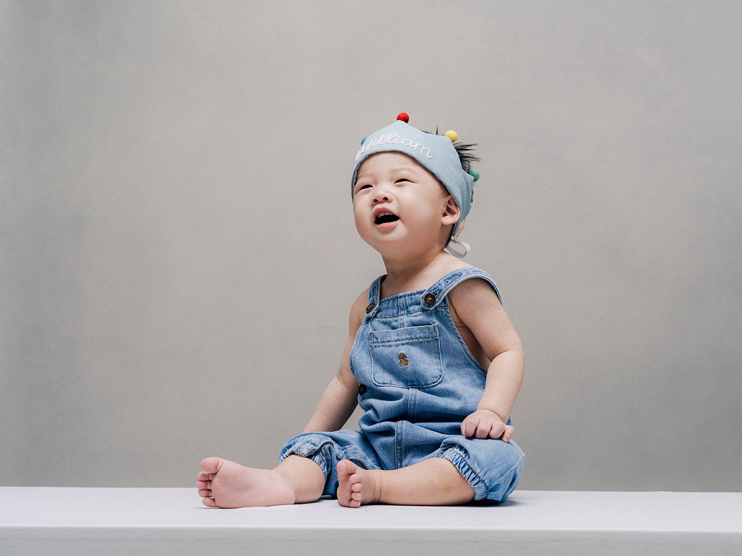 Baby wearing blue denim dress and white hat laughs while sitting on white surface against gray background.