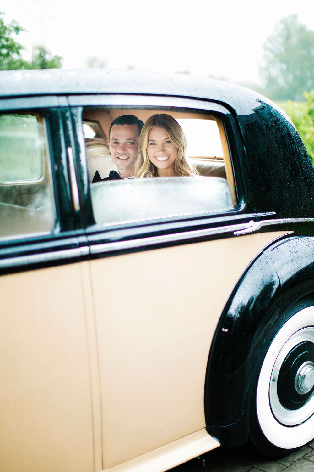 A couple smiles from inside a vintage black and cream colored car.