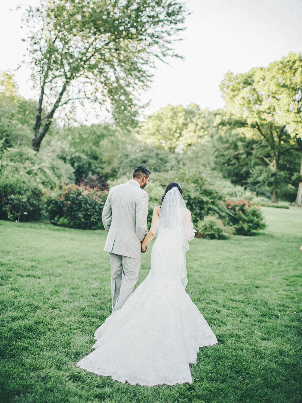 Newlyweds walk hand in hand through a lush green garden setting in elegant wedding attire.