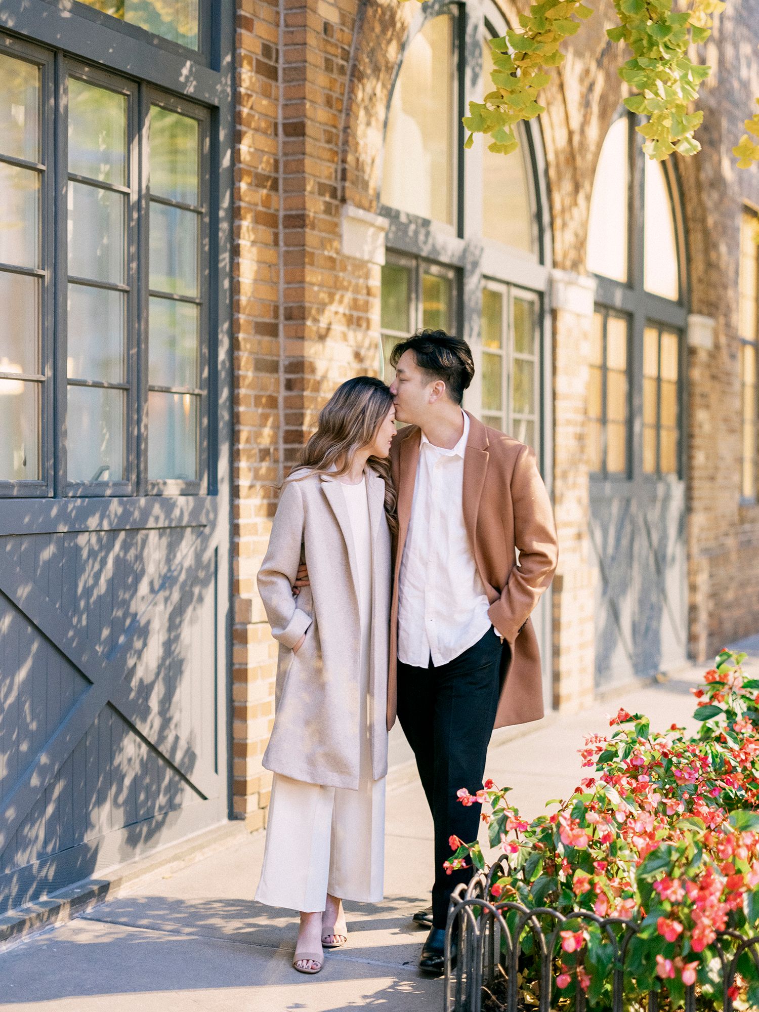 A couple strolls along a brick building with blooming flowers.