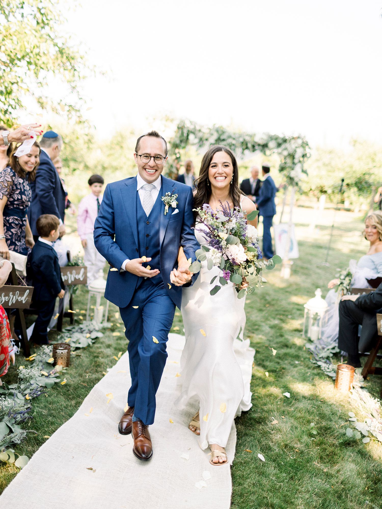 Newlyweds walk down aisle together holding bouquet at outdoor garden ceremony.