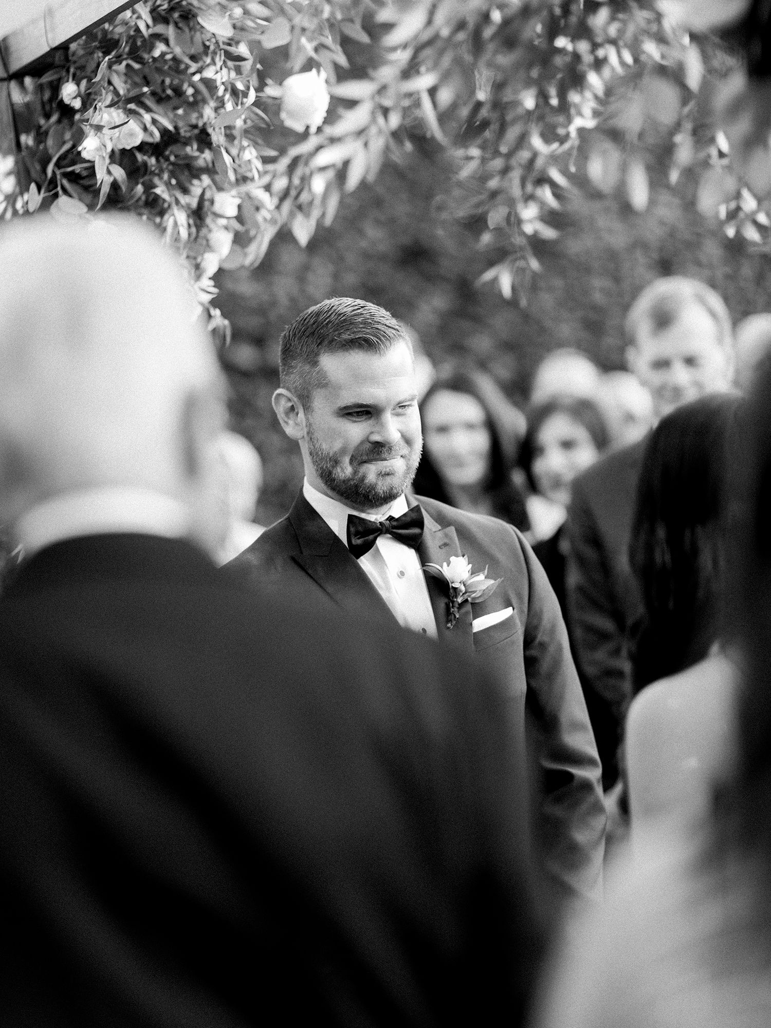 Wedding attendees in formal attire gathered beneath trees at an outdoor celebration.