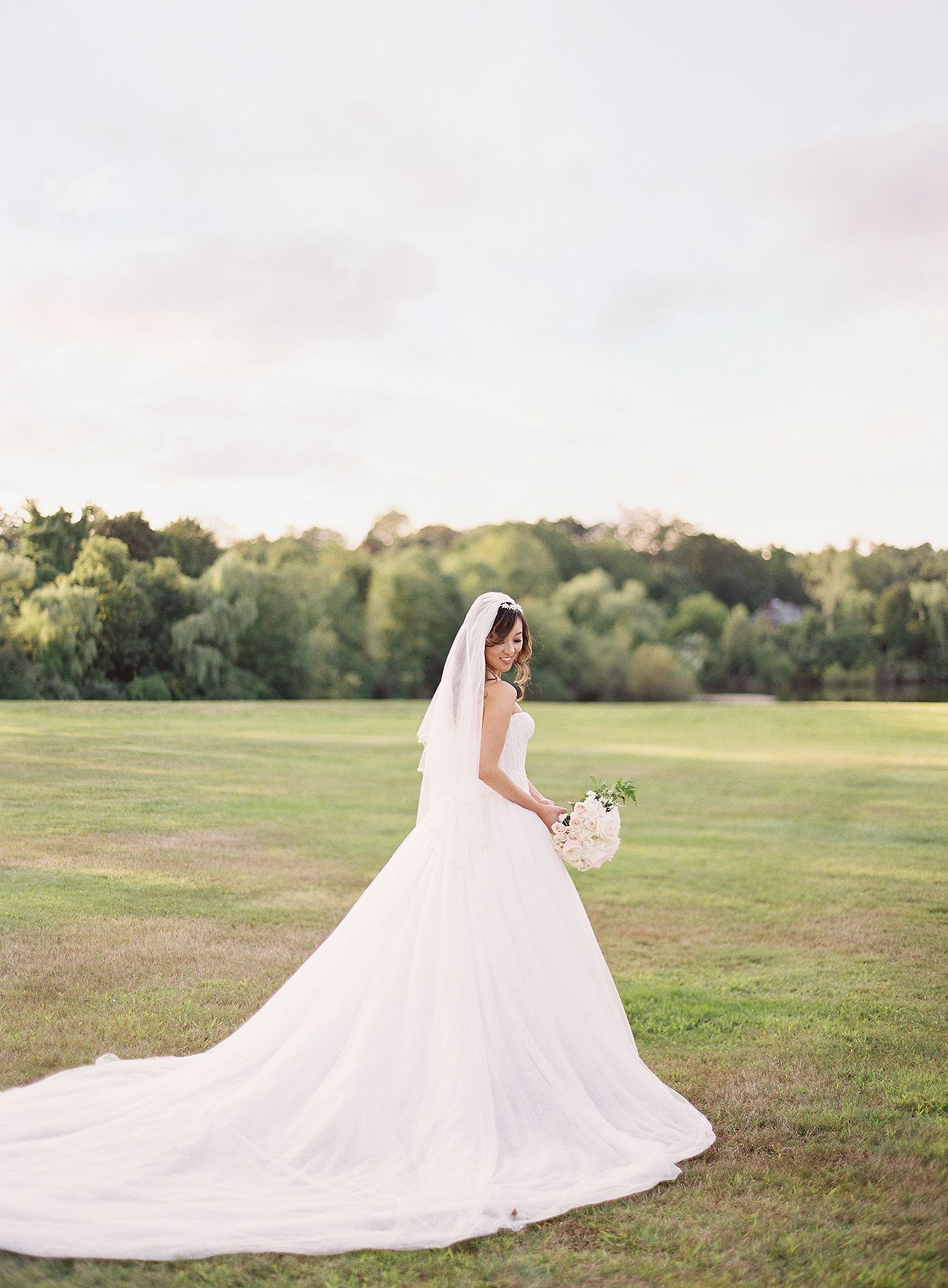 A flowing white wedding gown with a dramatic train spreads across an open grassy field at sunset. NYC wedding photographer.