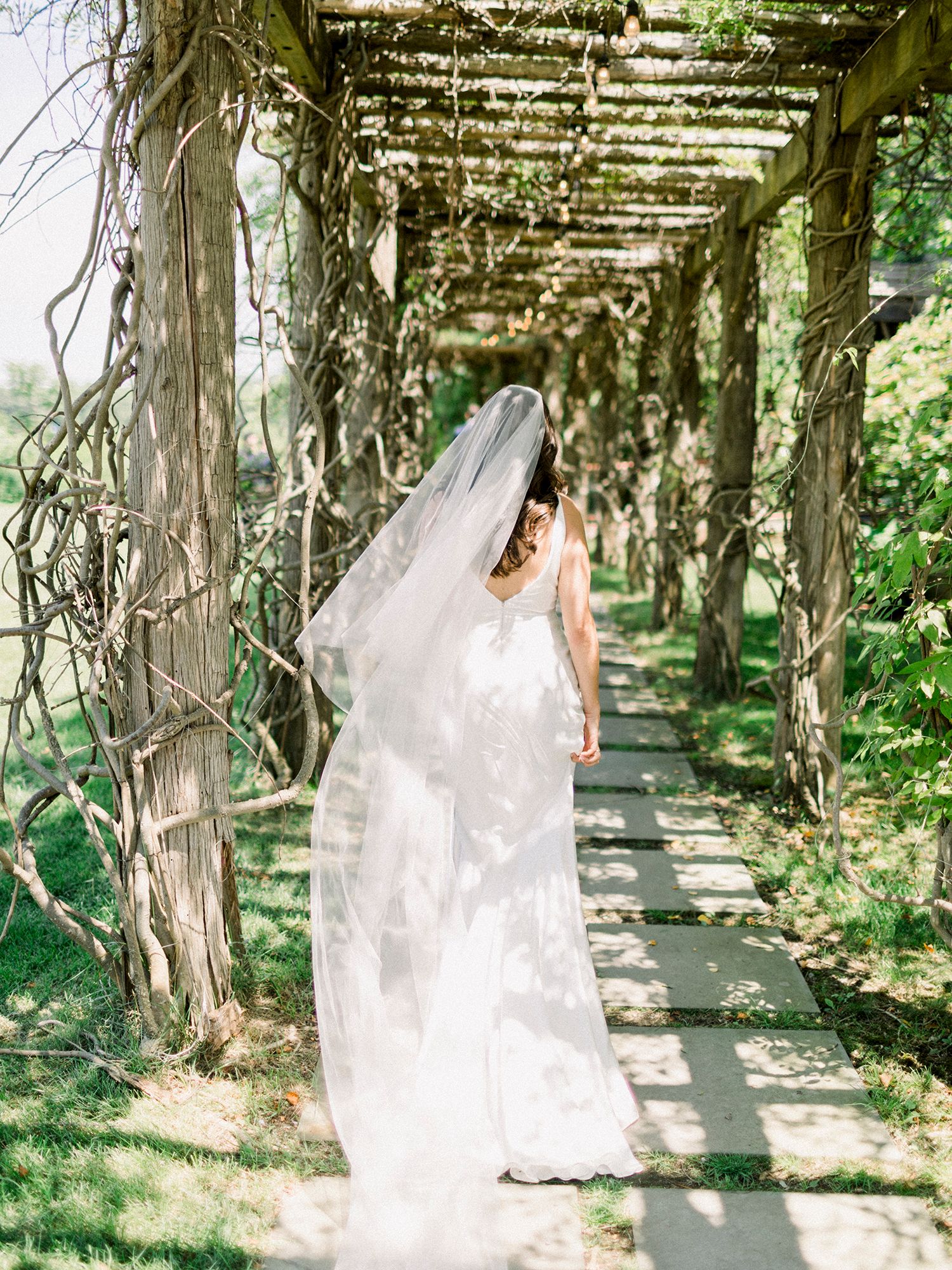 Bride in flowing white dress walking through a vine-covered garden pergola.