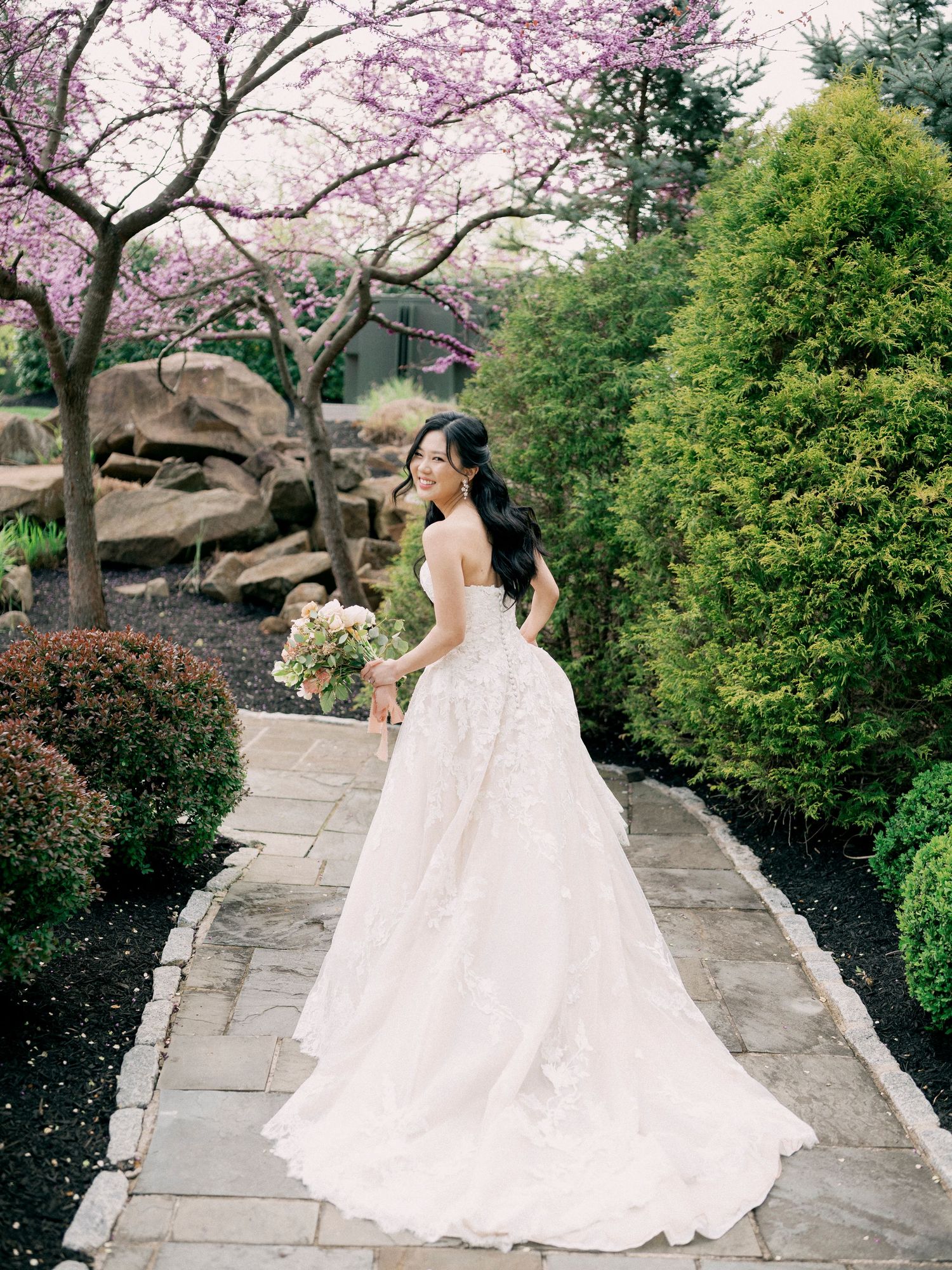 Wedding couple walking along a stone path under blooming cherry trees in a garden setting.