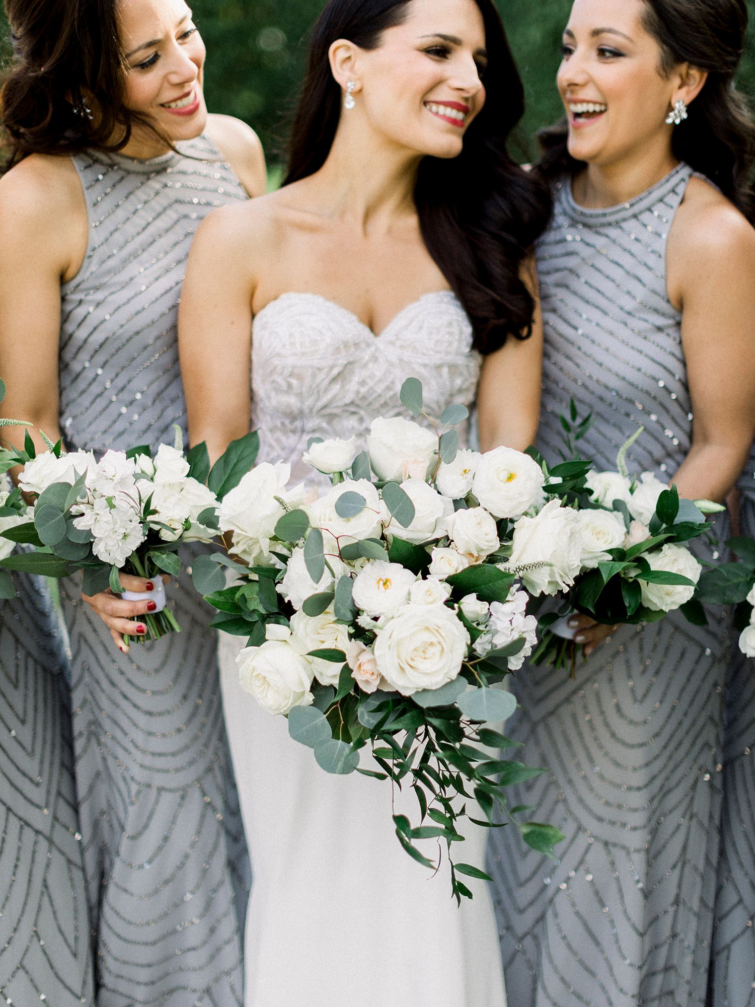 Bridesmaids in silver beaded dresses hold elegant white floral bouquets while smiling together.