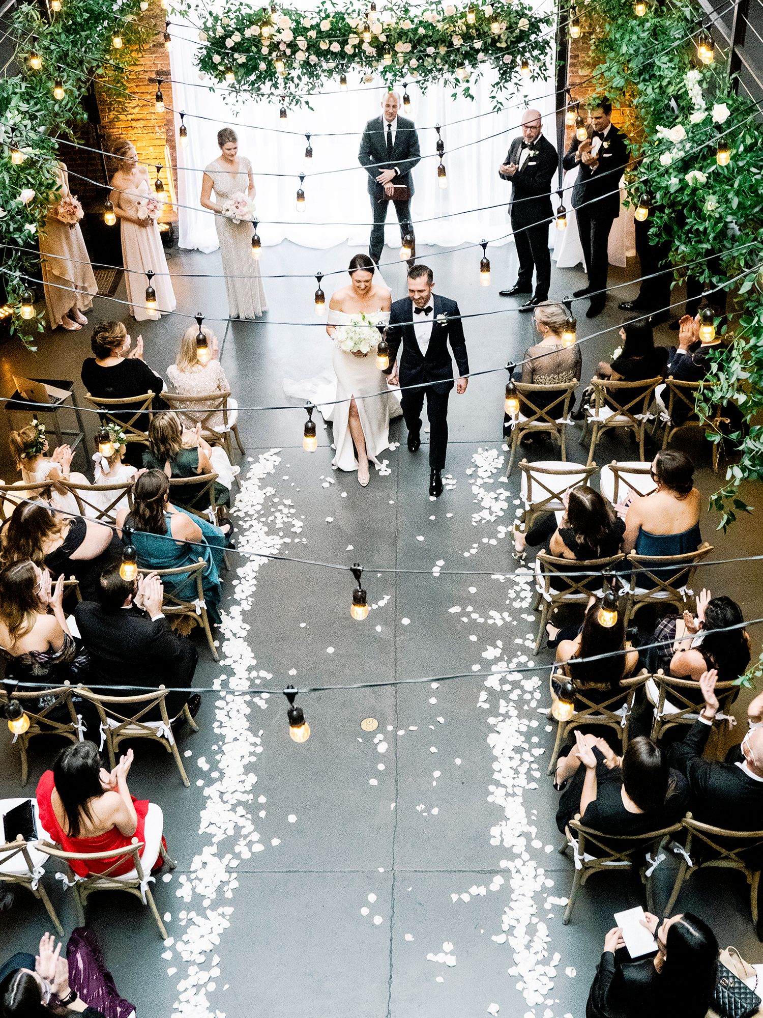 Wedding ceremony aisle decorated with greenery arch and string lights while guests watch from both sides. NYC wedding photographer.