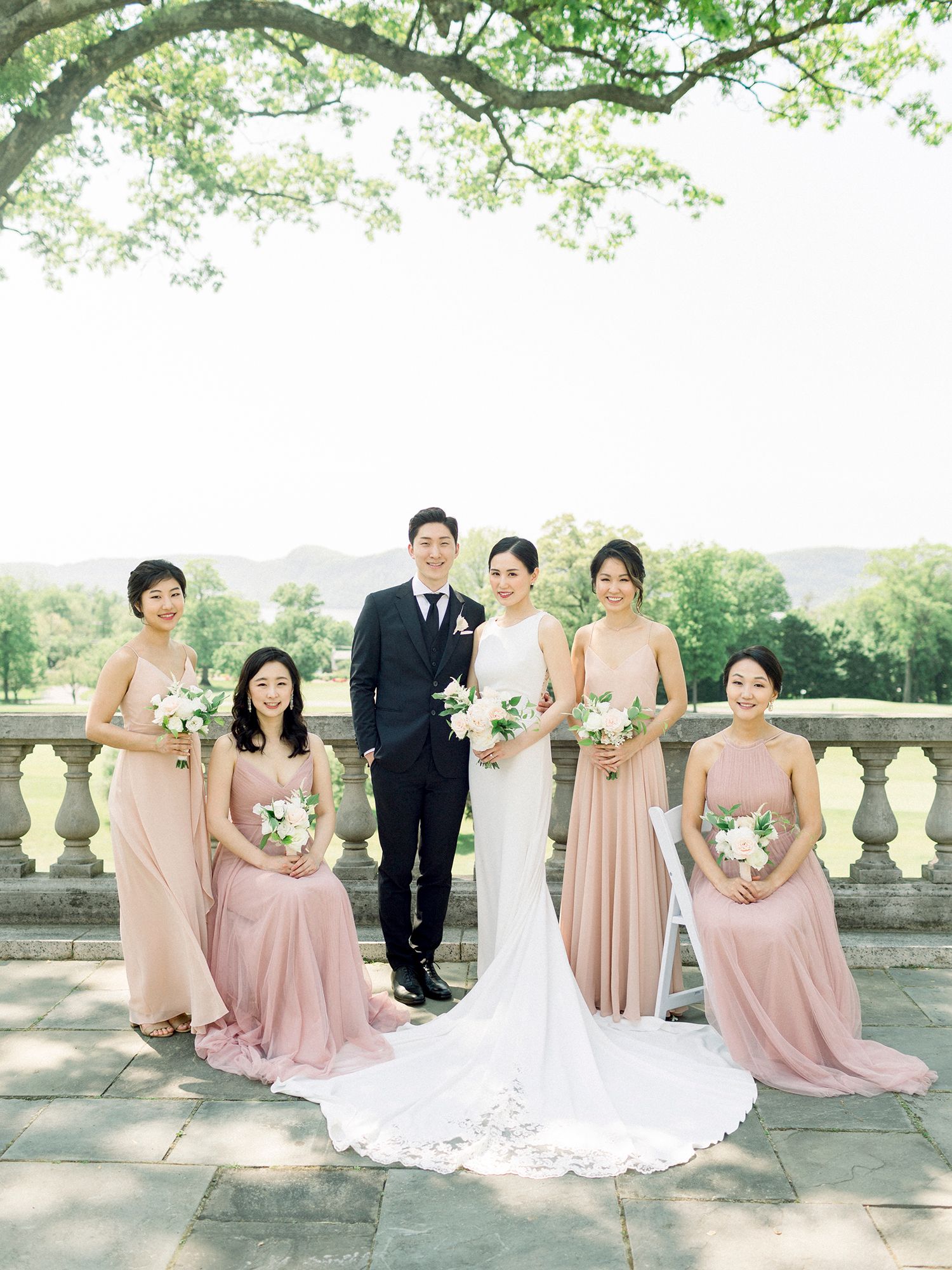 Wedding party in blush pink dresses poses on stone terrace under blooming spring trees.