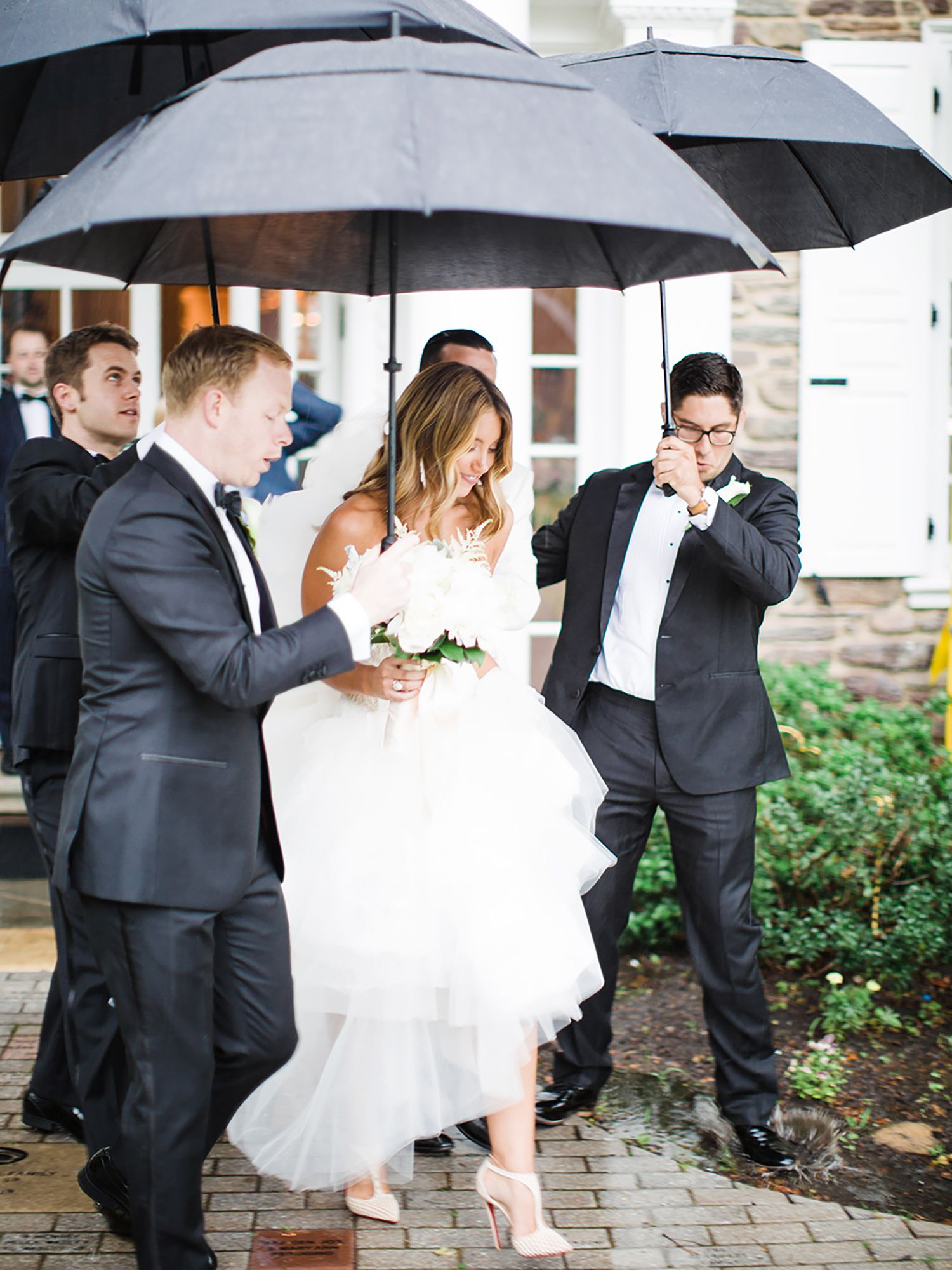 Wedding party poses playfully with black umbrellas on a rainy day outside.