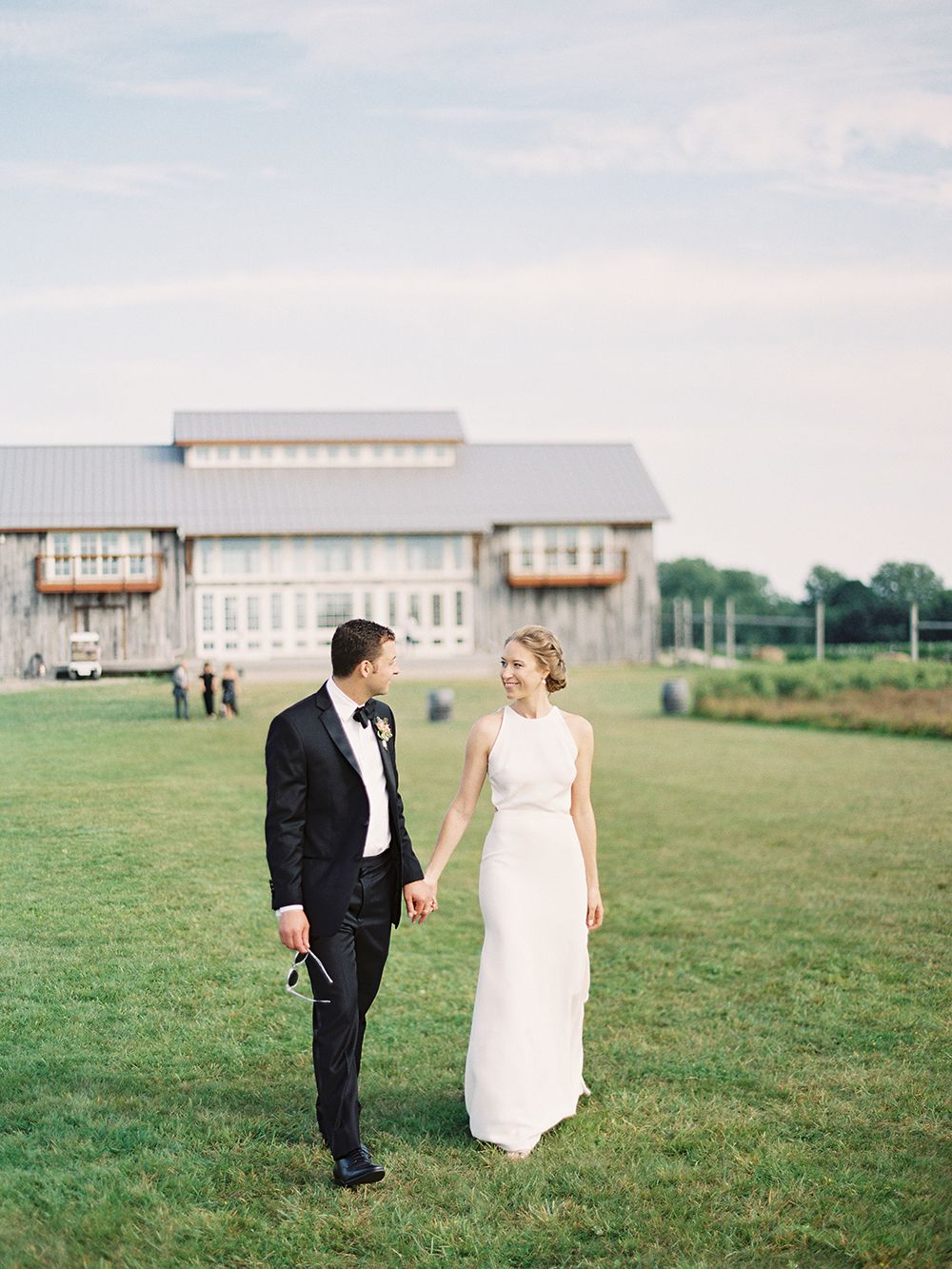 A couple walks across a green lawn with a rustic barn venue in the background. NYC wedding photographer.