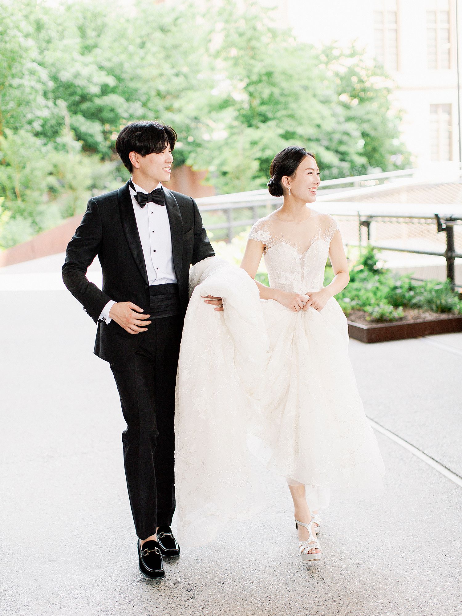 A couple in formal wedding attire playfully walks and dances together on an outdoor pathway. NYC wedding photographer.