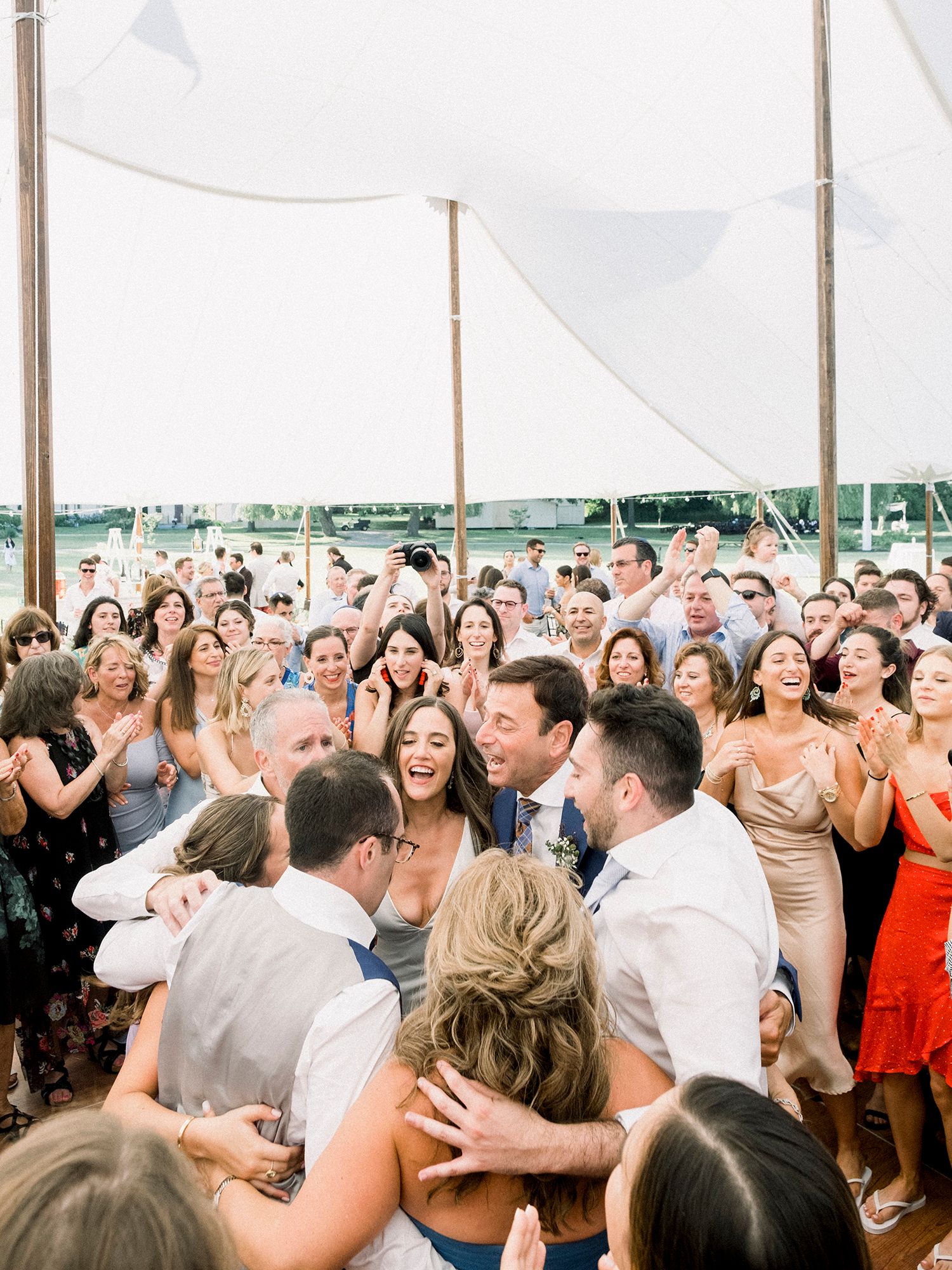 A large crowd of wedding guests dance and celebrate together under a white tent.