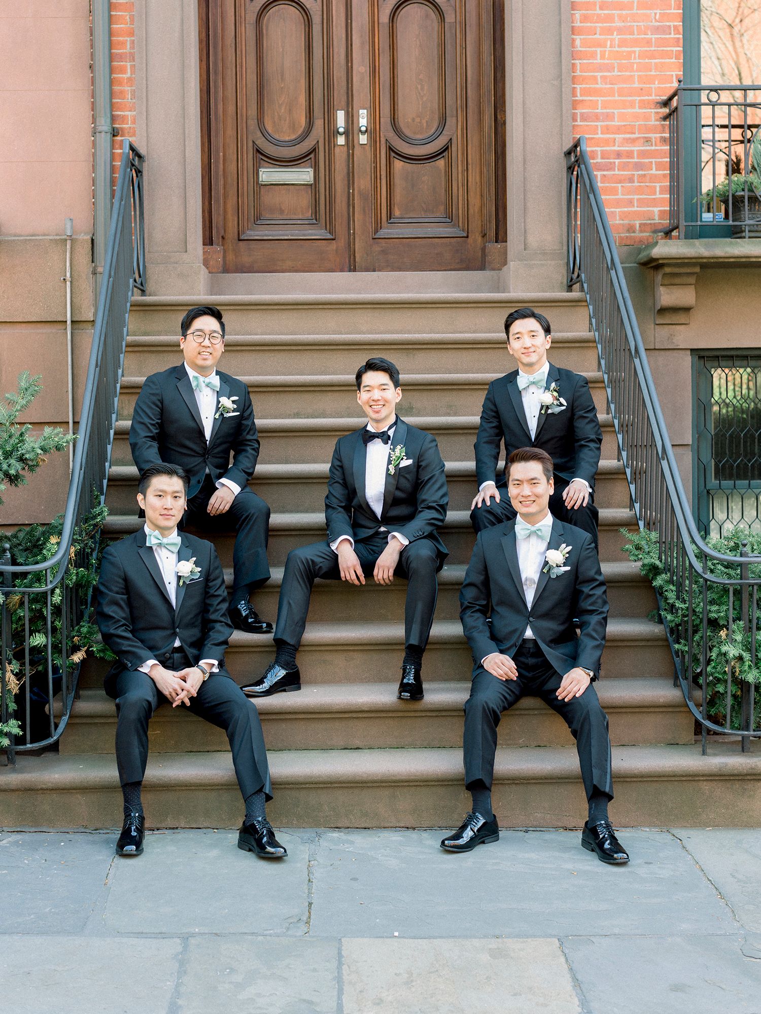 Groomsmen in black tuxedos sit casually on brick building steps.